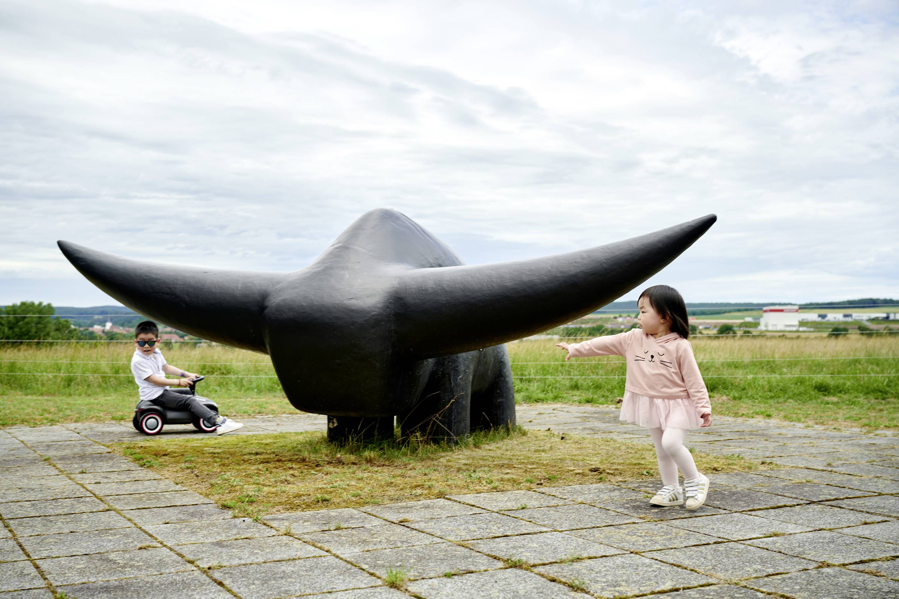 Zwei Kinder spielen neben einer großen schwarzen Stier-Skulptur mit ausgebreiteten Hörnern auf einer Wiese unter bewölktem Himmel.