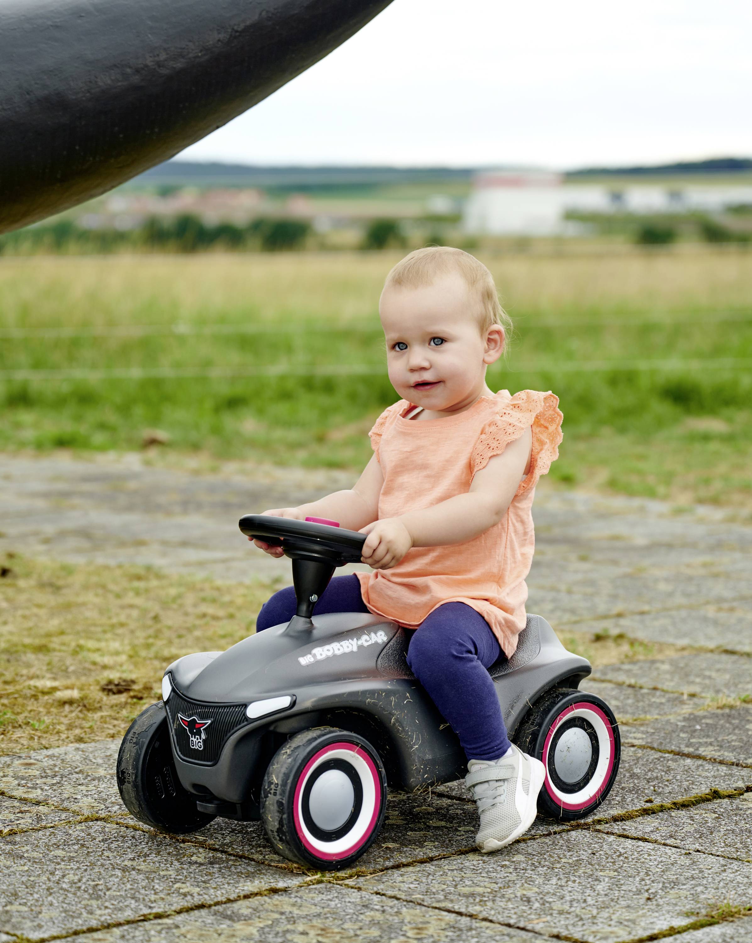 Ein Kleinkind sitzt auf einem kleinen, schwarzen Kinderfahrzeug im Freien. Es trägt ein orangefarbenes Shirt und schaut in die Kamera.