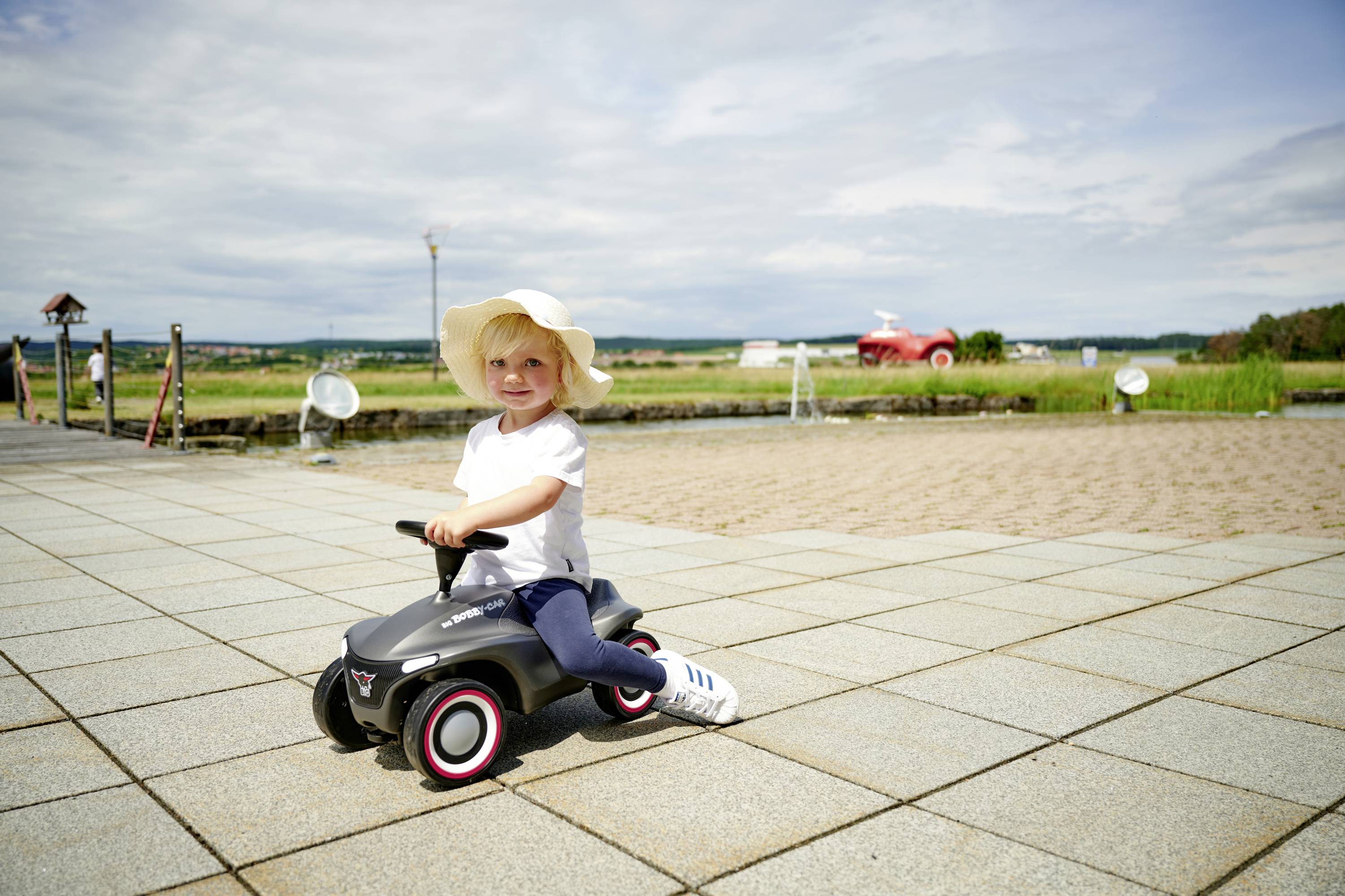 Ein kleines Kind mit Sonnenhut fährt auf einem Spielzeugauto auf einer gefliesten Terrasse. Im Hintergrund ist eine Grünfläche zu sehen.