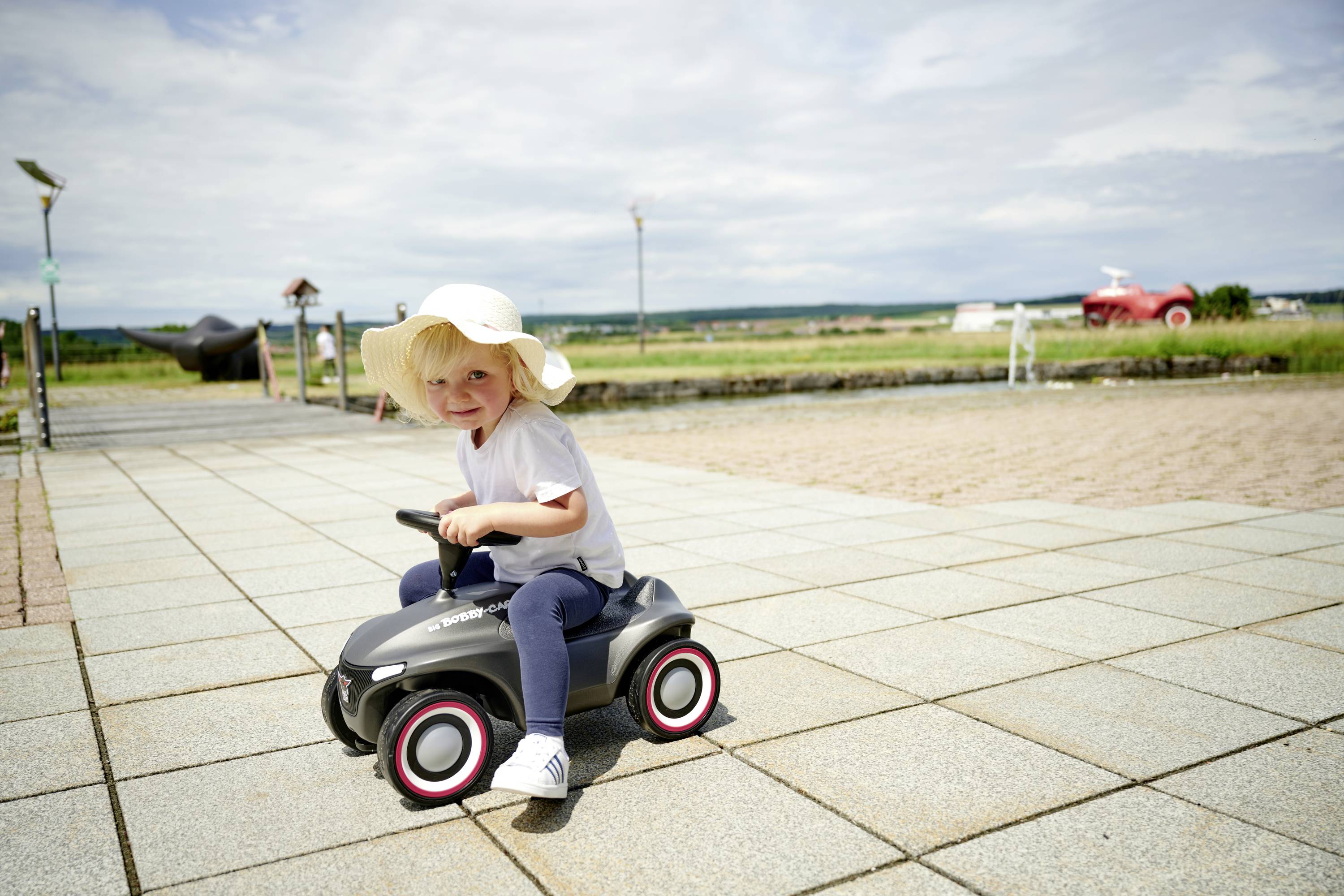 Ein kleines Kind mit einem weißen Hut sitzt fröhlich auf einem Spielzeugauto draußen auf einem gepflasterten Platz bei sonnigem Wetter.