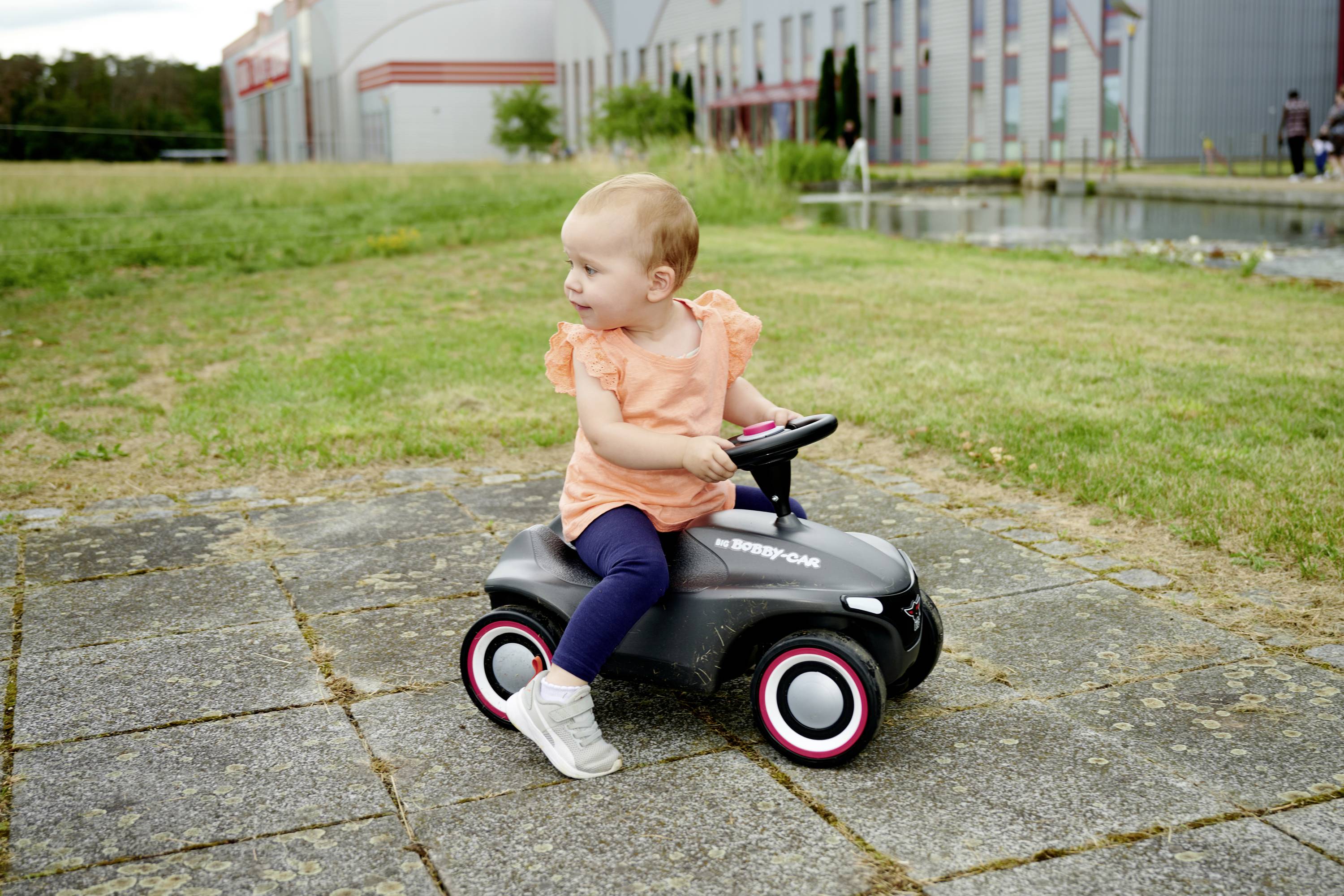 Ein Kleinkind sitzt auf einem Spielzeugauto im Freien, umgeben von Rasen und einem Gebäude im Hintergrund. Das Kind trägt ein orangefarbenes Oberteil.