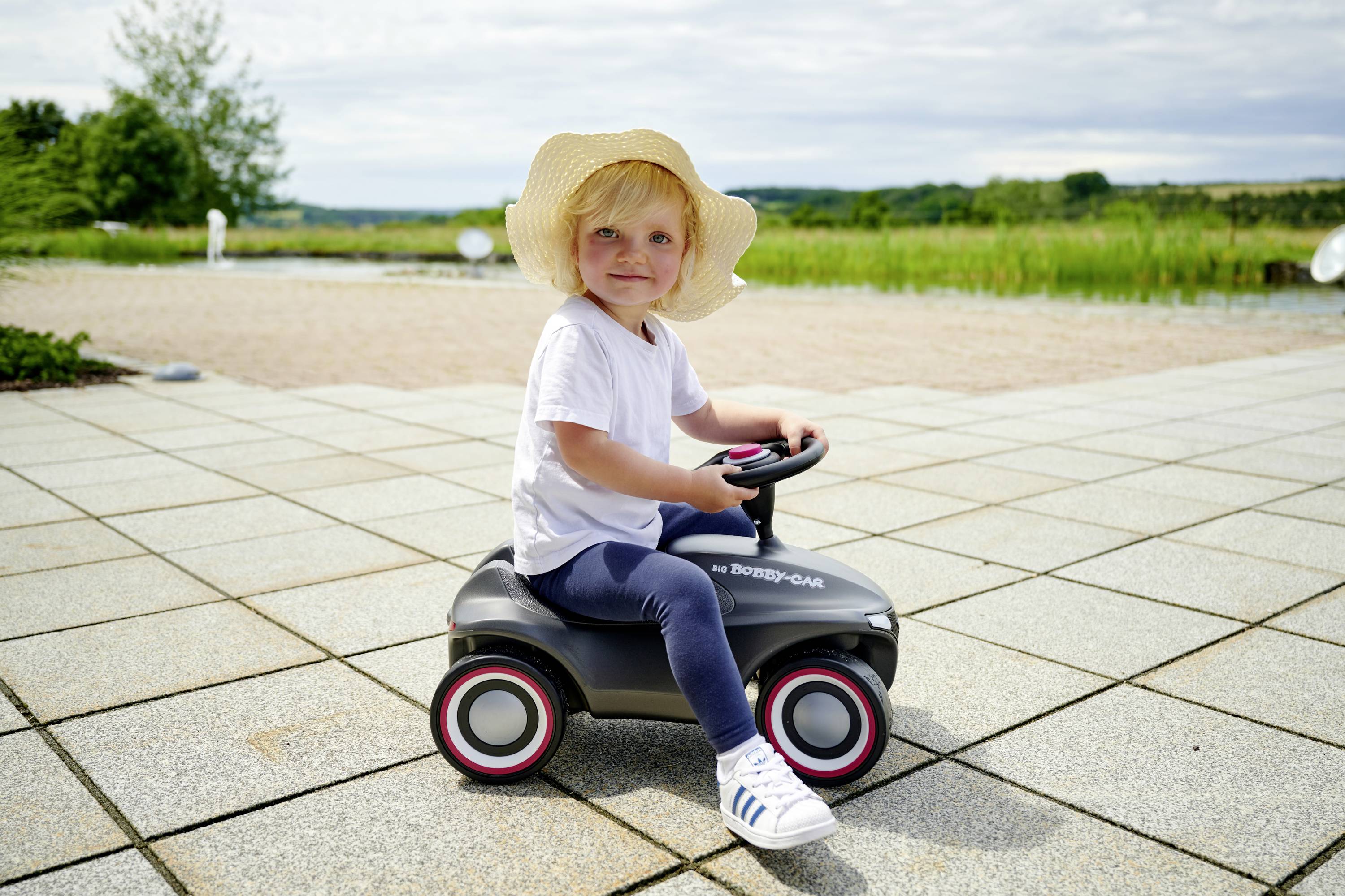 Kleinkind mit Strohhut sitzt auf einem Spielzeugauto draußen auf einer Terrasse, im Hintergrund eine grüne Wiese und ein bewölkter Himmel.