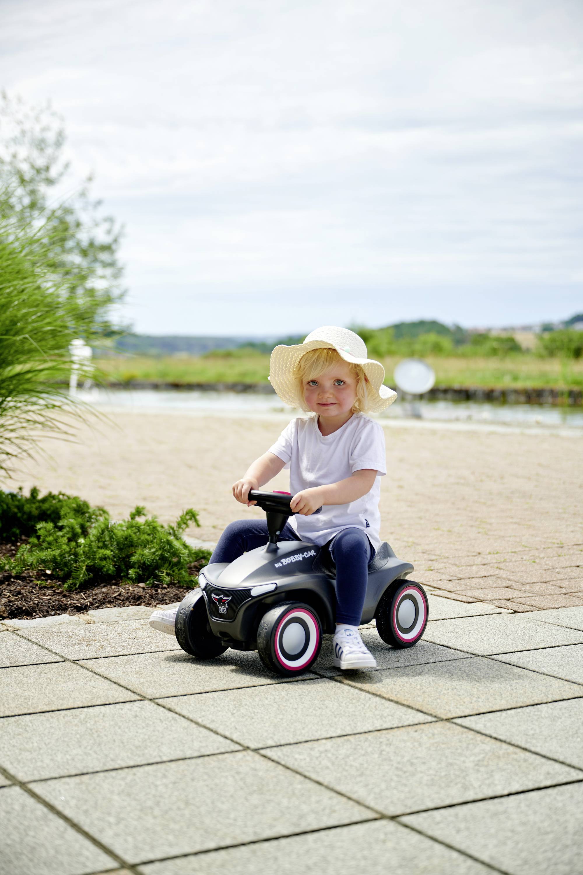 Ein kleines Kind mit Strohhut sitzt auf einem schwarzen Spielzeugauto im Freien auf einer gepflasterten Fläche. Im Hintergrund Natur.