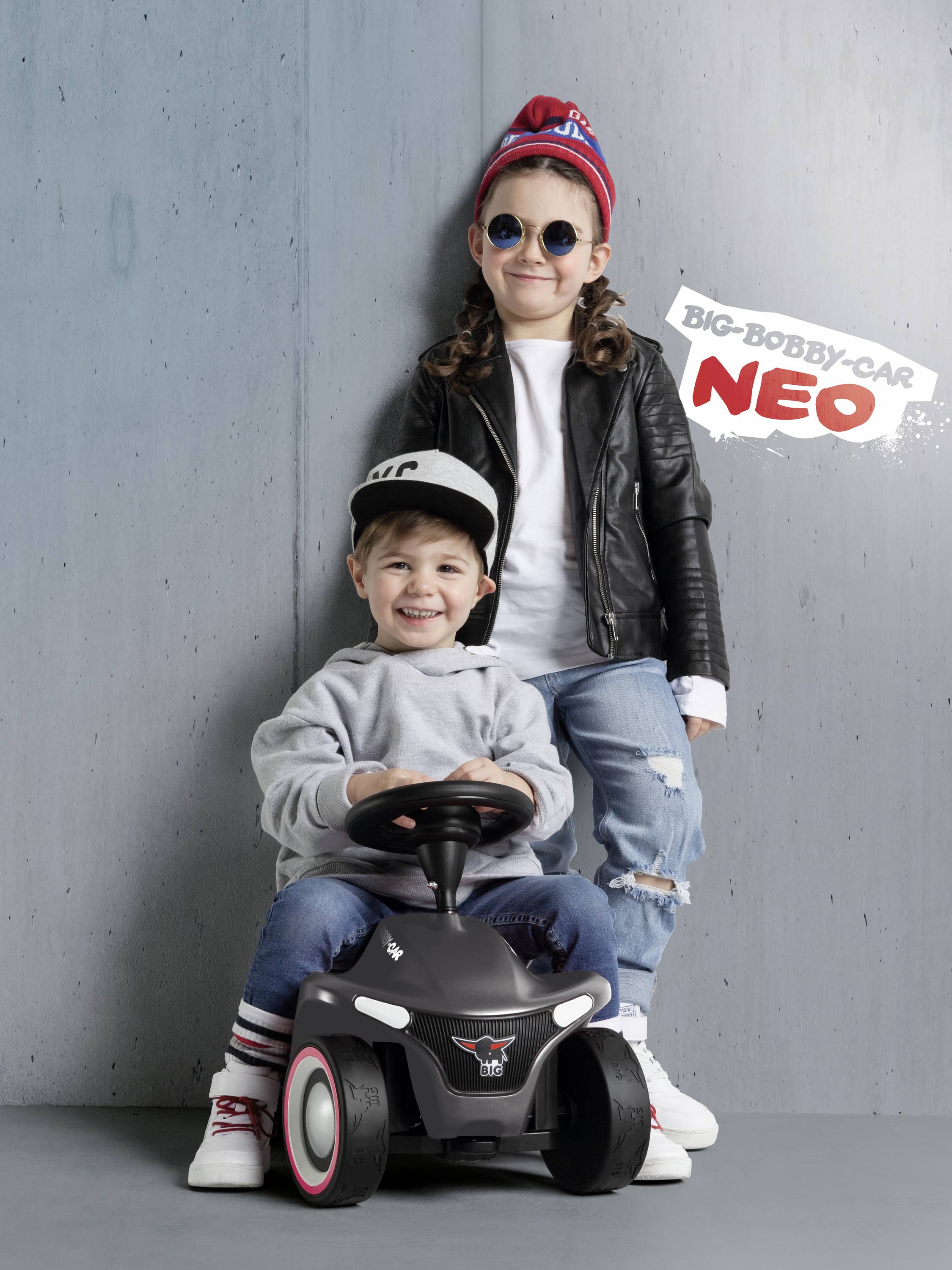 Zwei Kinder posieren fröhlich mit einem kleinen Kinderfahrzeug. Beide tragen modische Kleidung und Mützen.
