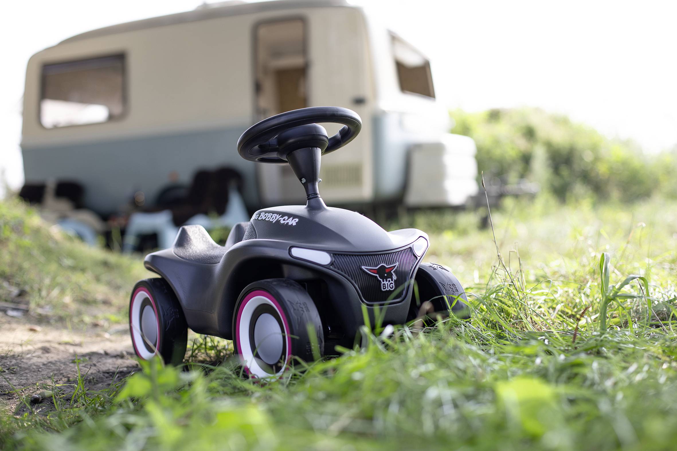 Ein schwarzes Kinderfahrzeug steht auf grünem Gras, im Hintergrund ist ein unscharfer Wohnwagen zu sehen.