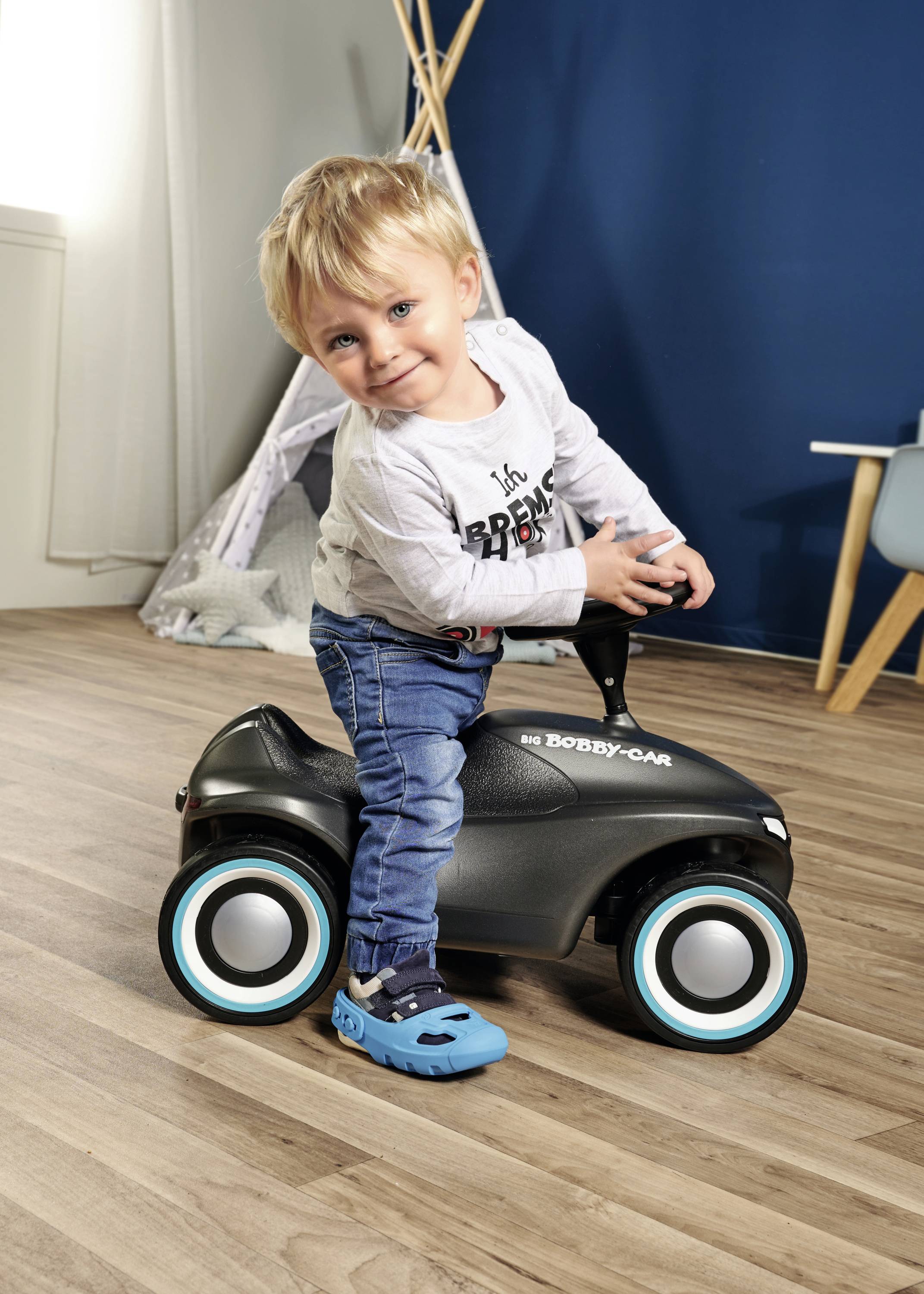 Ein Kleinkind fährt lächelnd auf einem schwarzen Bobby-Car in einem gemütlichen Raum mit Holzfußboden und blauen Wänden.