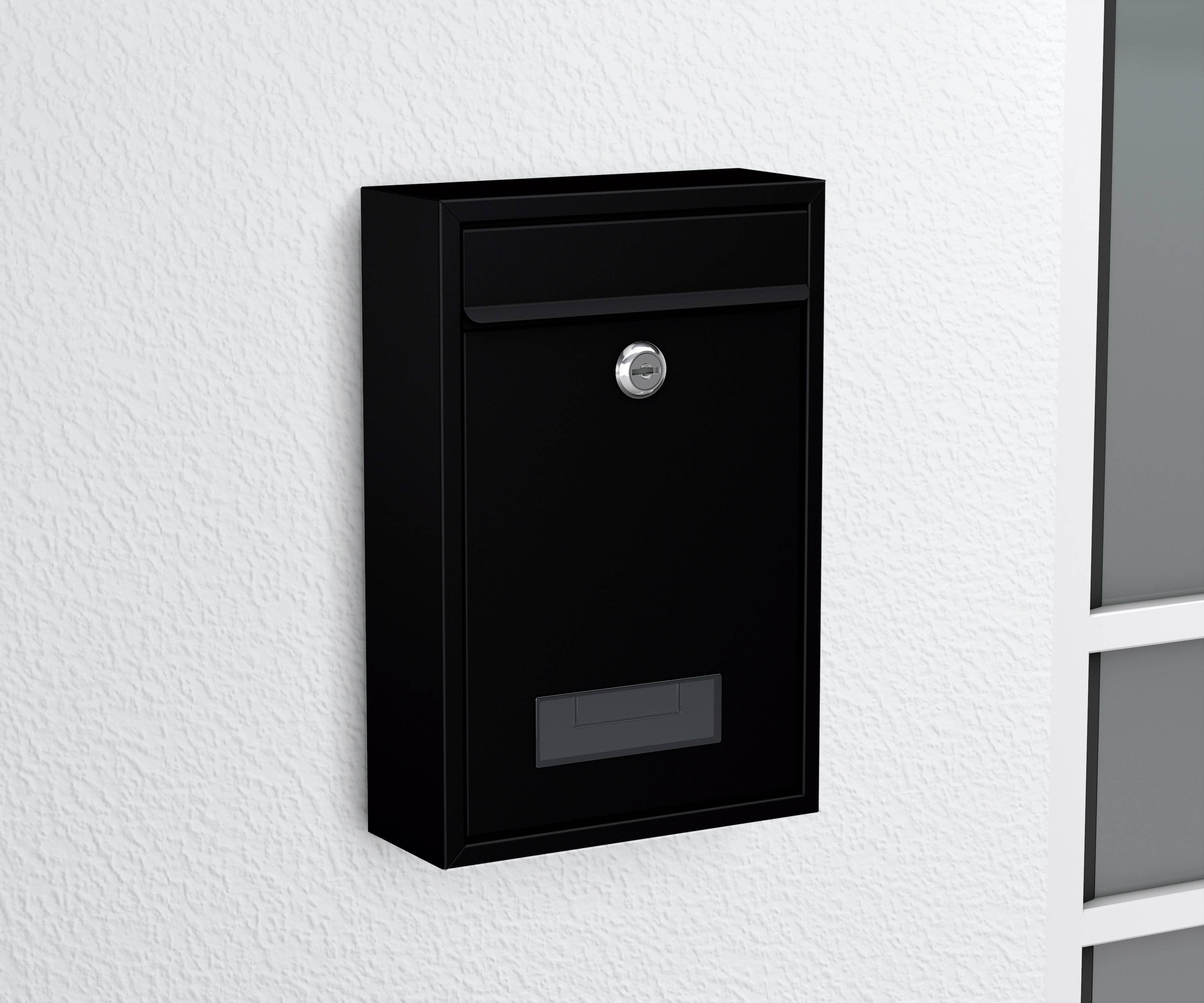 Ein schwarzer Briefkasten an einer weißen Wand, rechts neben einer Tür mit Glaseinsatz.