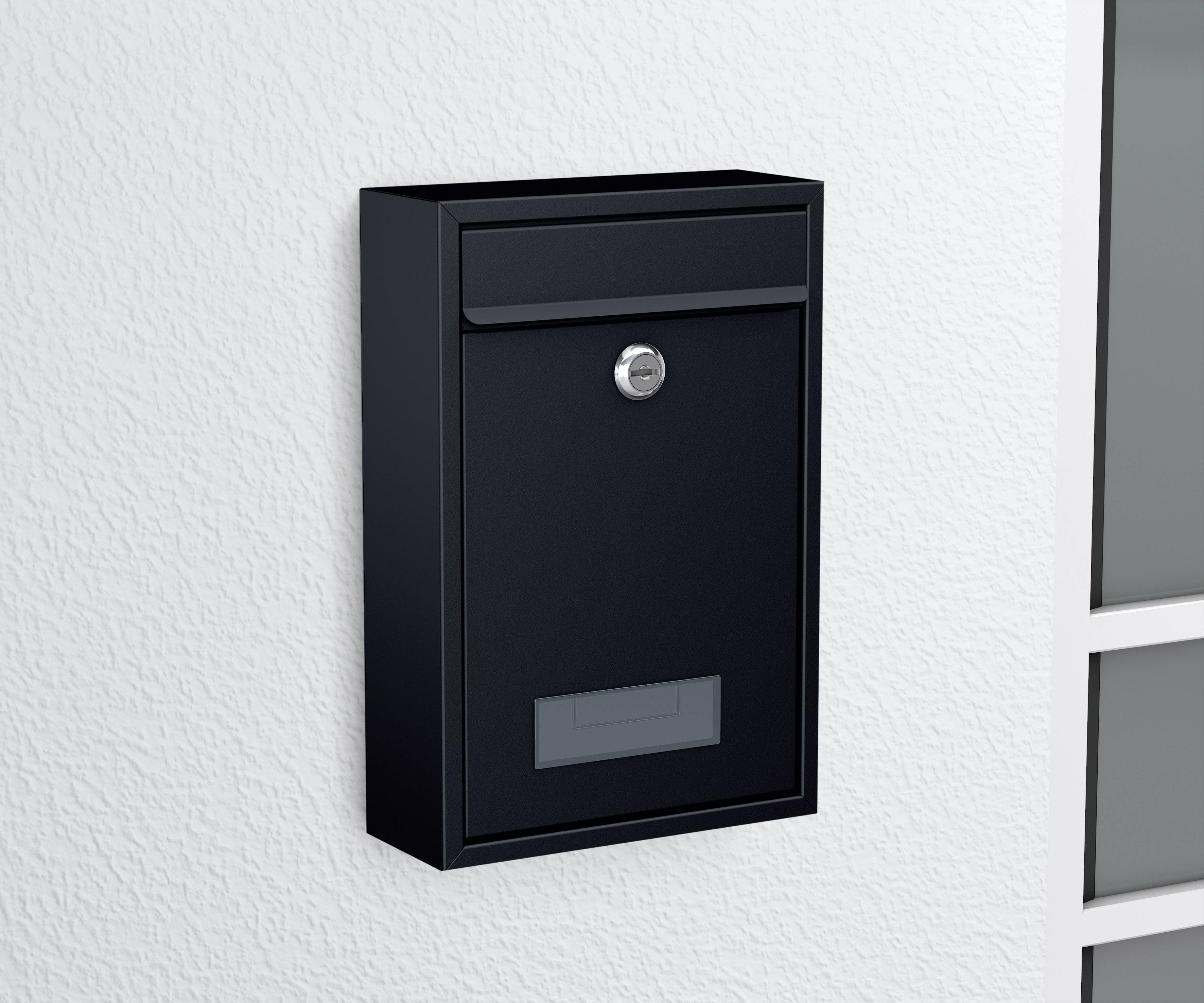 Ein schwarzer Briefkasten an einer weißen Wand, abgeschlossen mit einem silbernen Schloss, neben einem Fenster mit vier Scheiben.