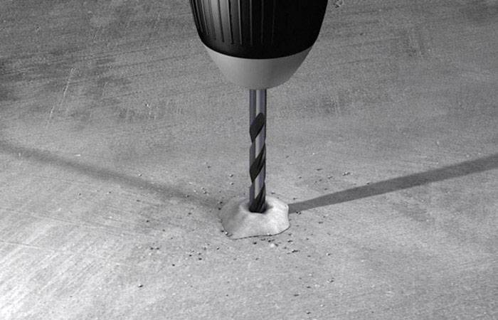 Eine Bohrmaschine bohrt in Beton, Staub bildet sich um das Bohrloch. Die Bohrmaschine ist schwarz und grau, die Umgebung grau.