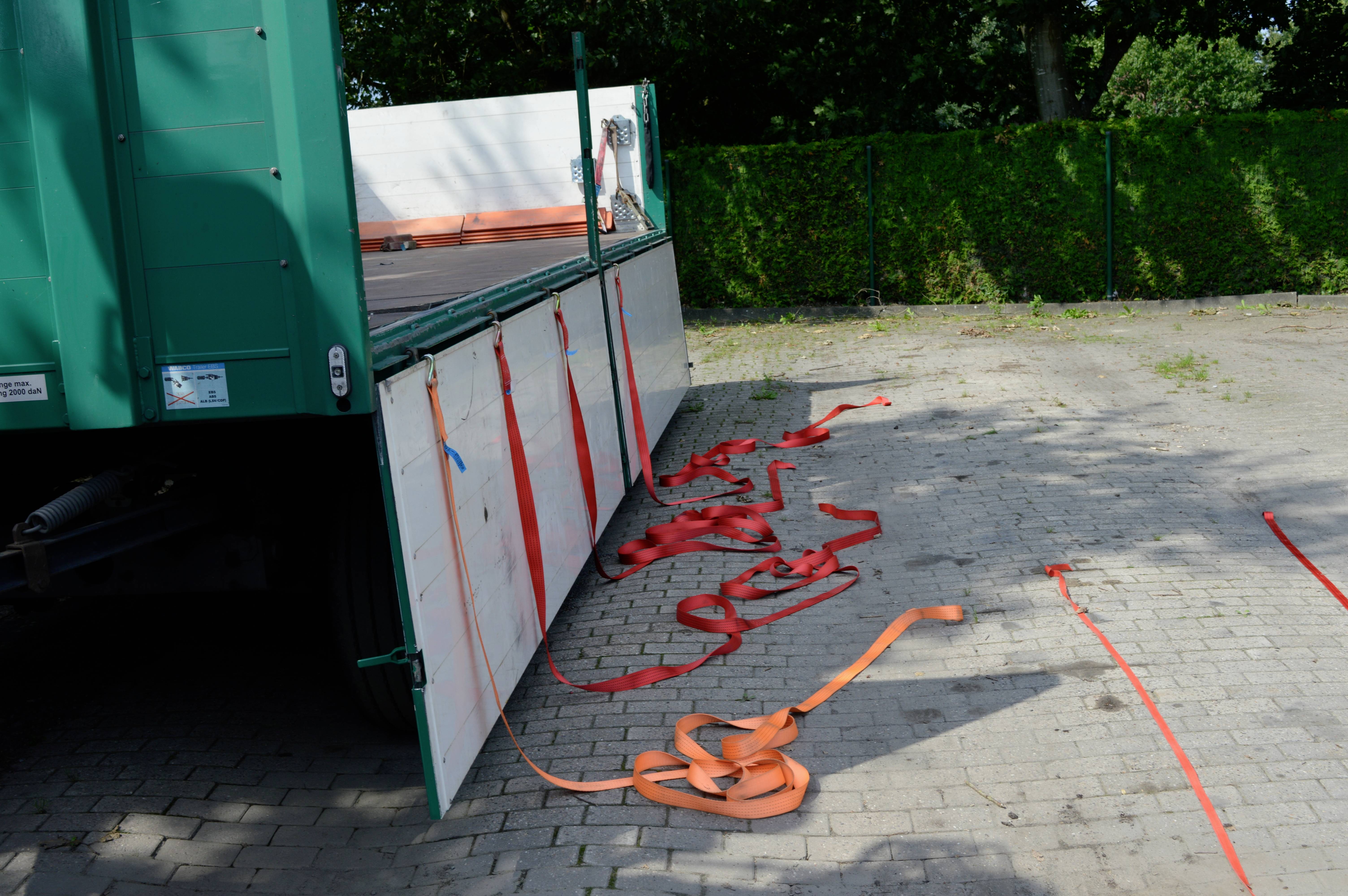 Ein LKW mit offenen Seitenwänden auf einem gepflasterten Hof. Mehrere rote und orangefarbene Gurte liegen auf dem Boden.