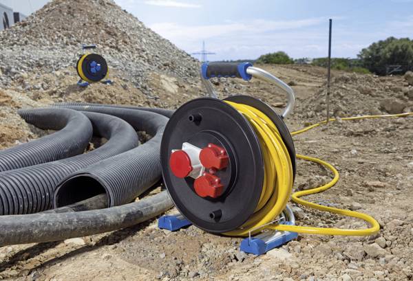 Ein gelbes Kabel auf einer Kabeltrommel auf einer Baustelle. Im Hintergrund sind Schutthaufen und große schwarze Röhren zu sehen.