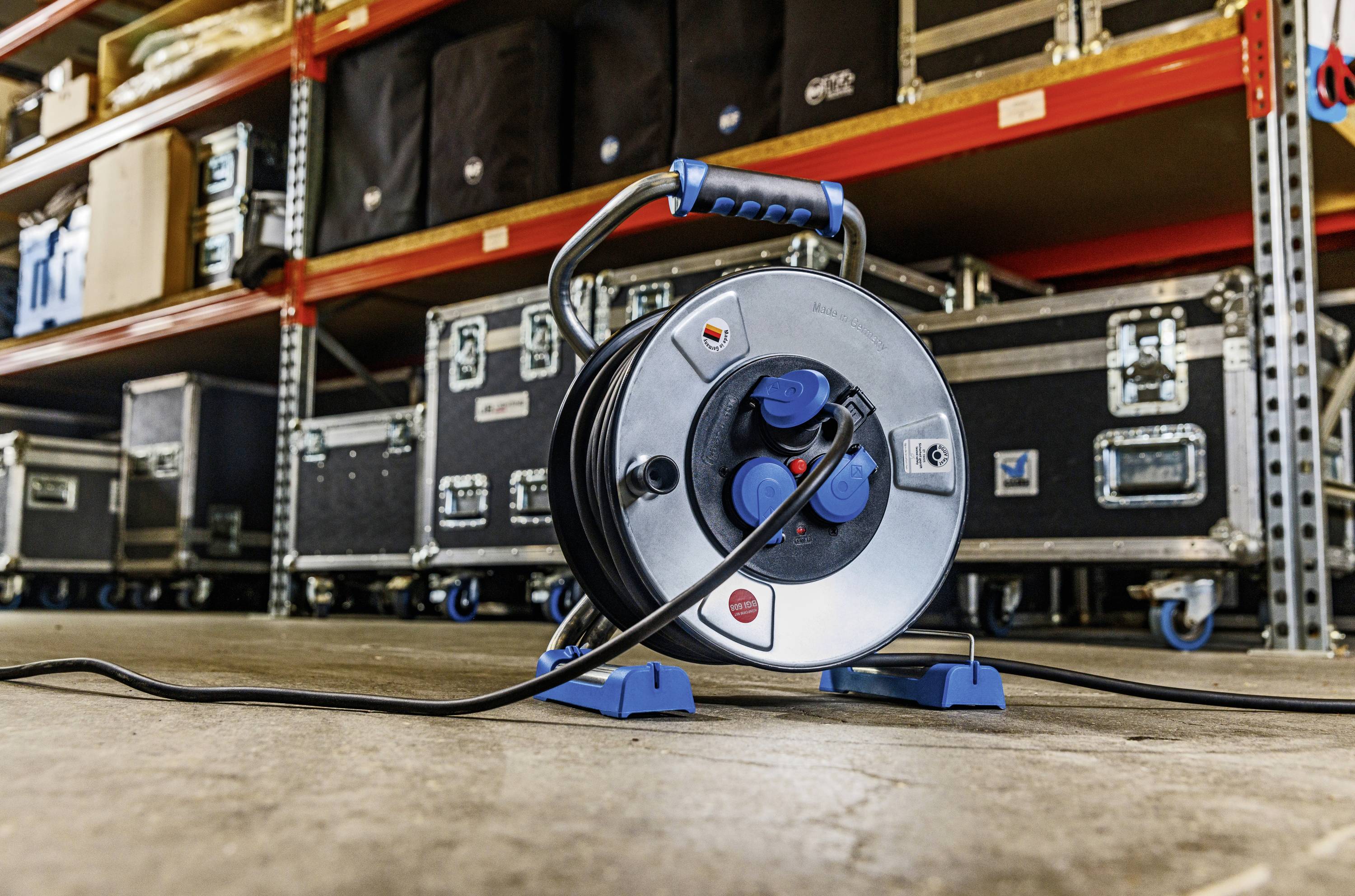 Eine Kabeltrommel steht auf dem Boden in einem Lagerraum mit Regalen voller Ausrüstung und Transportkoffern im Hintergrund.