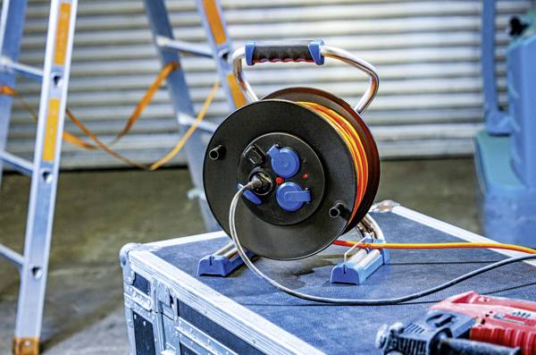 Eine Kabeltrommel mit orangem Verlängerungskabel steht auf einem schwarzen Kasten. Im Hintergrund sind eine Leiter und Metallwand sichtbar.