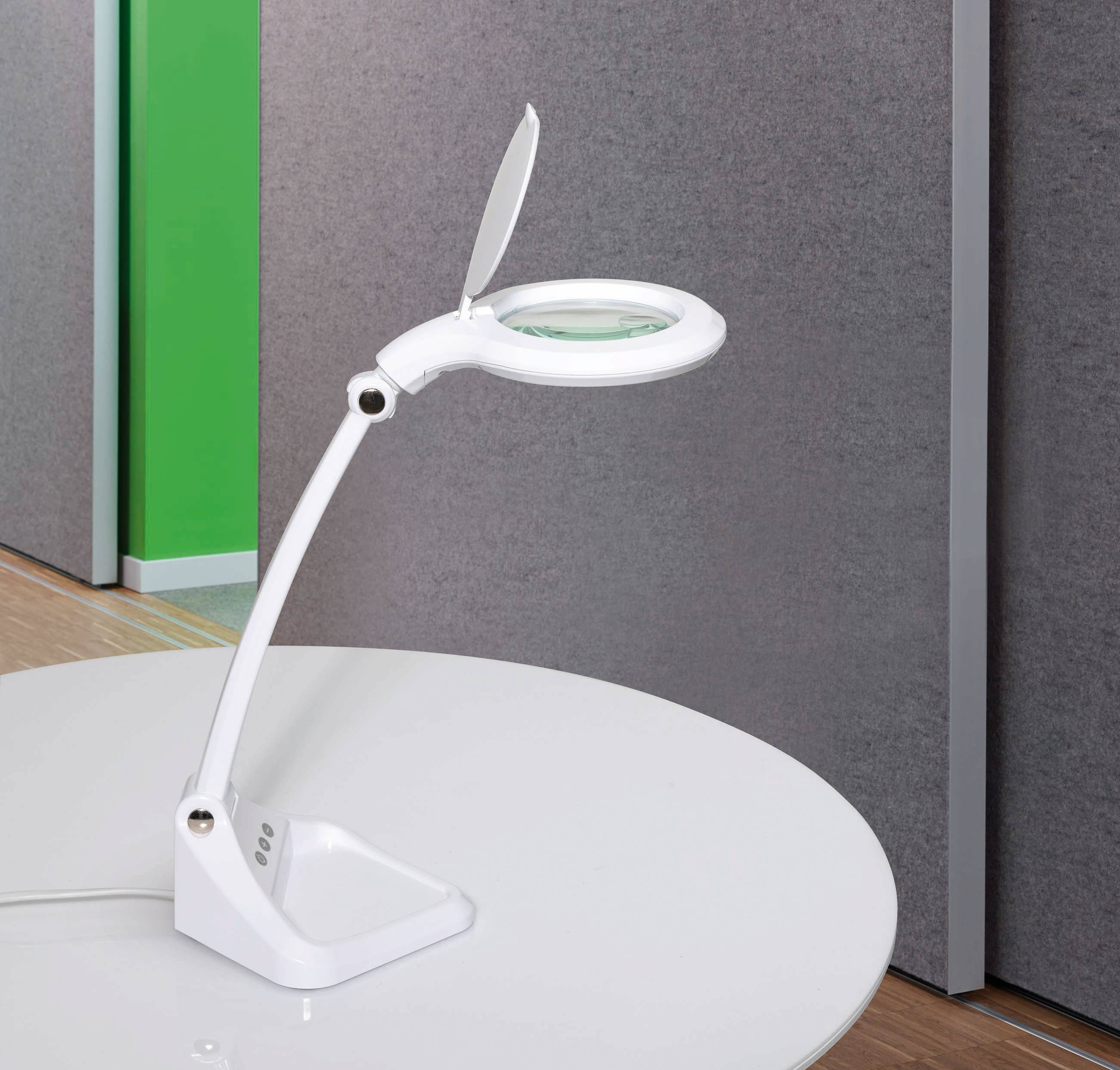 Eine weiße Tischlampe mit verstellbarem Arm und Lupenfunktion steht auf einem weißen Tisch vor einer grau-grünen Wand.