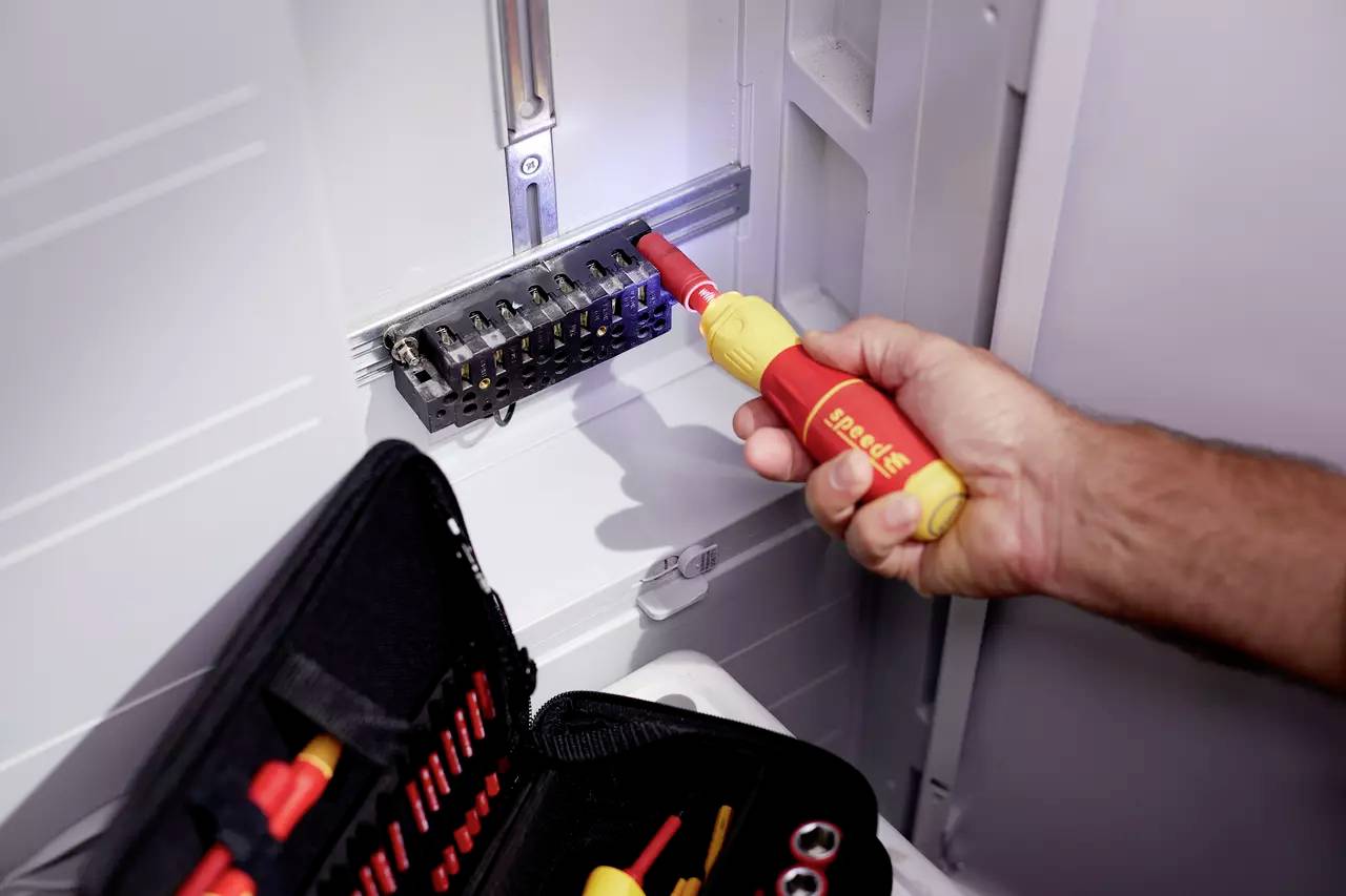 Eine Hand verwendet einen isolierten Schraubendreher, um an einer elektrischen Schalttafel zu arbeiten. Ein Werkzeugkoffer liegt daneben.