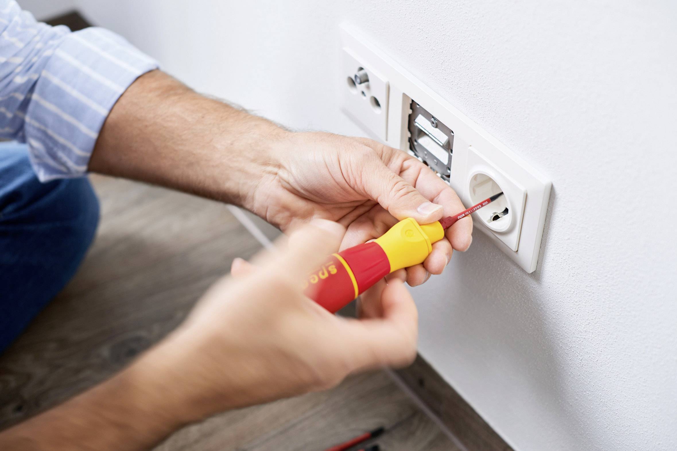 Eine Person repariert eine Wandsteckdose mit einem Schraubenzieher. Die Schrauben werden justiert. Es handelt sich um elektrische Arbeiten.