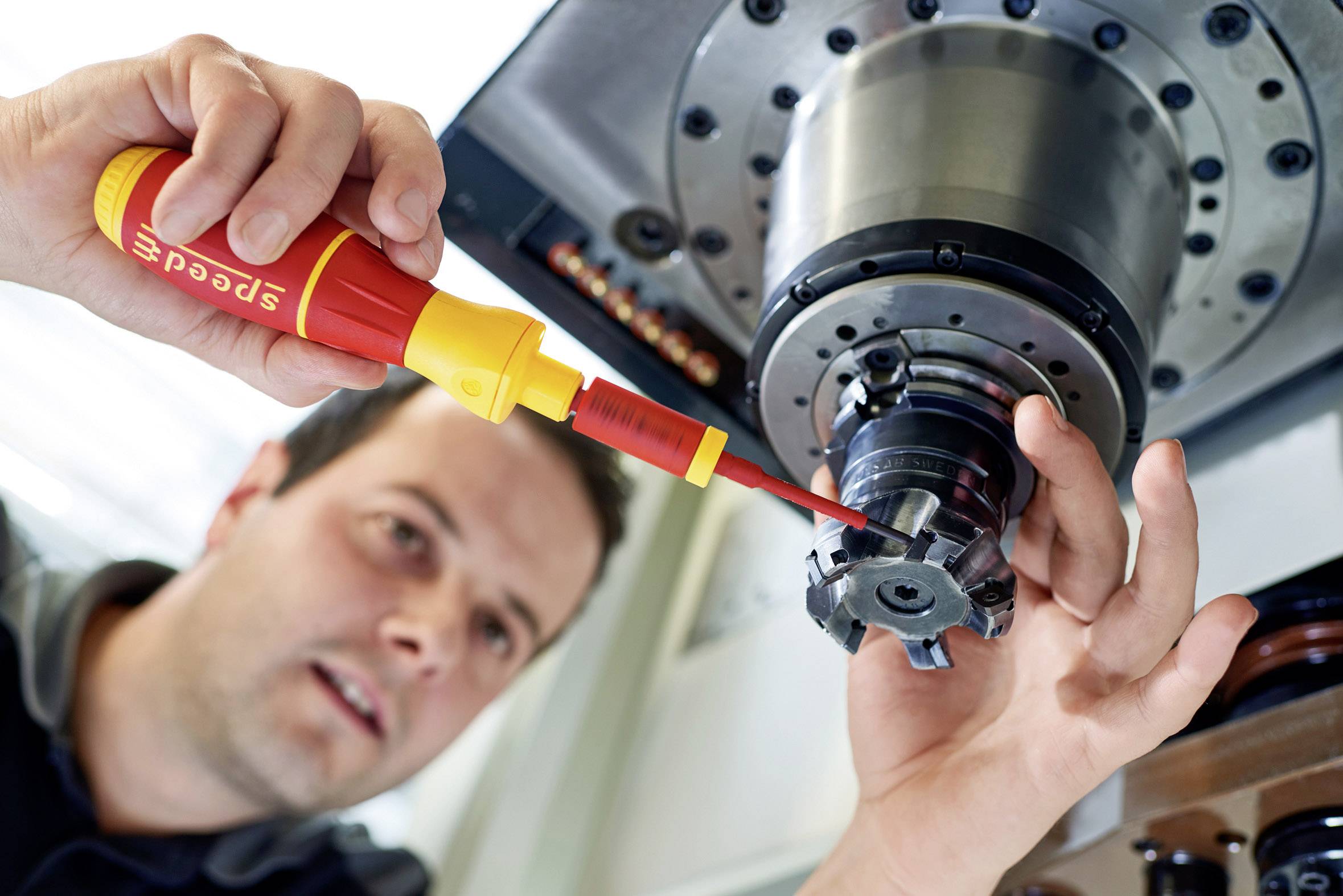 Eine Person justiert mit einem gelben Schraubendreher einen Metallbohrer an einer großen Maschine. Fokus liegt auf präziser Handarbeit.