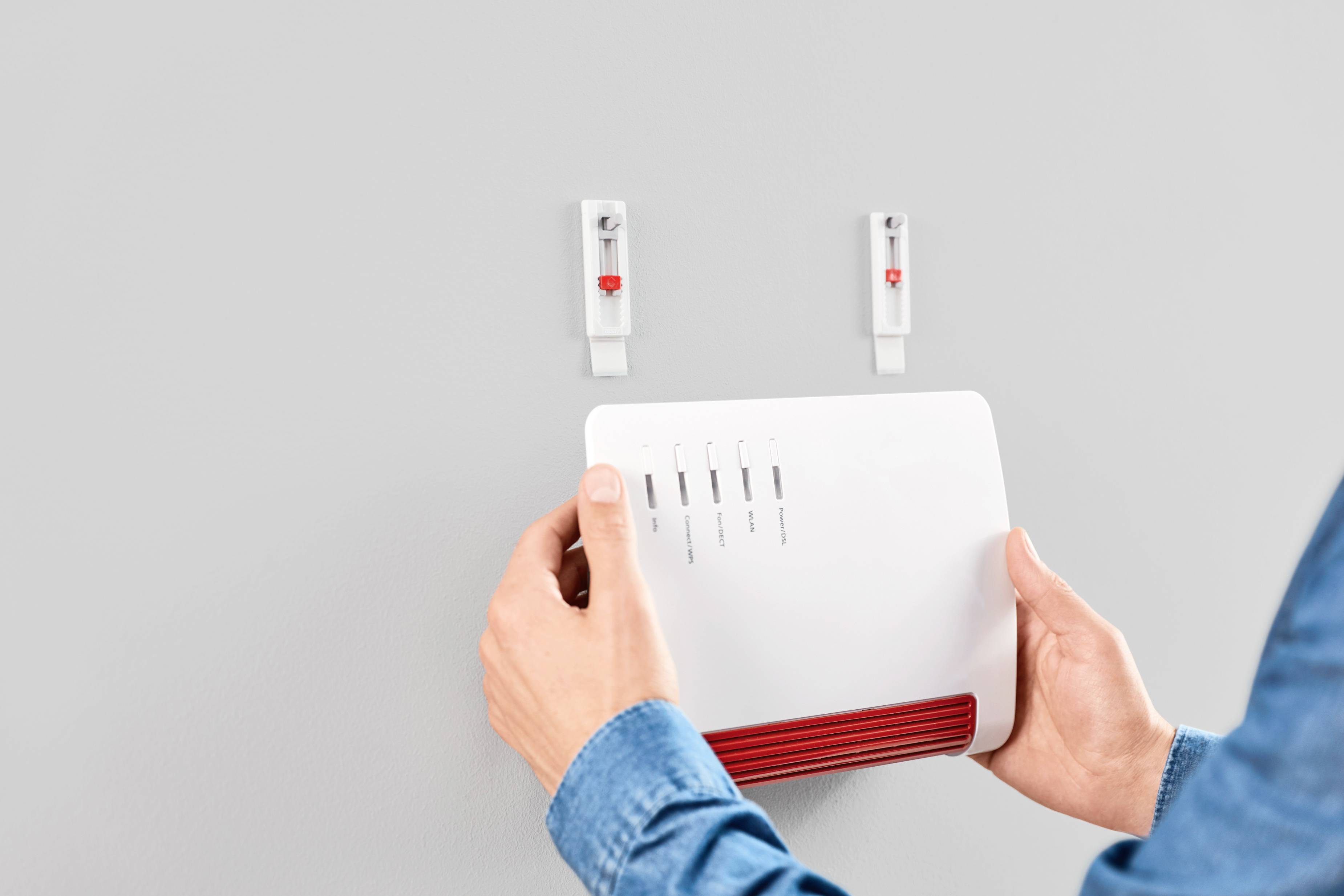 Person befestigt einen weißen WLAN-Router an der Wand. Der Router zeigt Signalstärke- und Verbindungssymbole. Zwei leere Haken sind sichtbar.