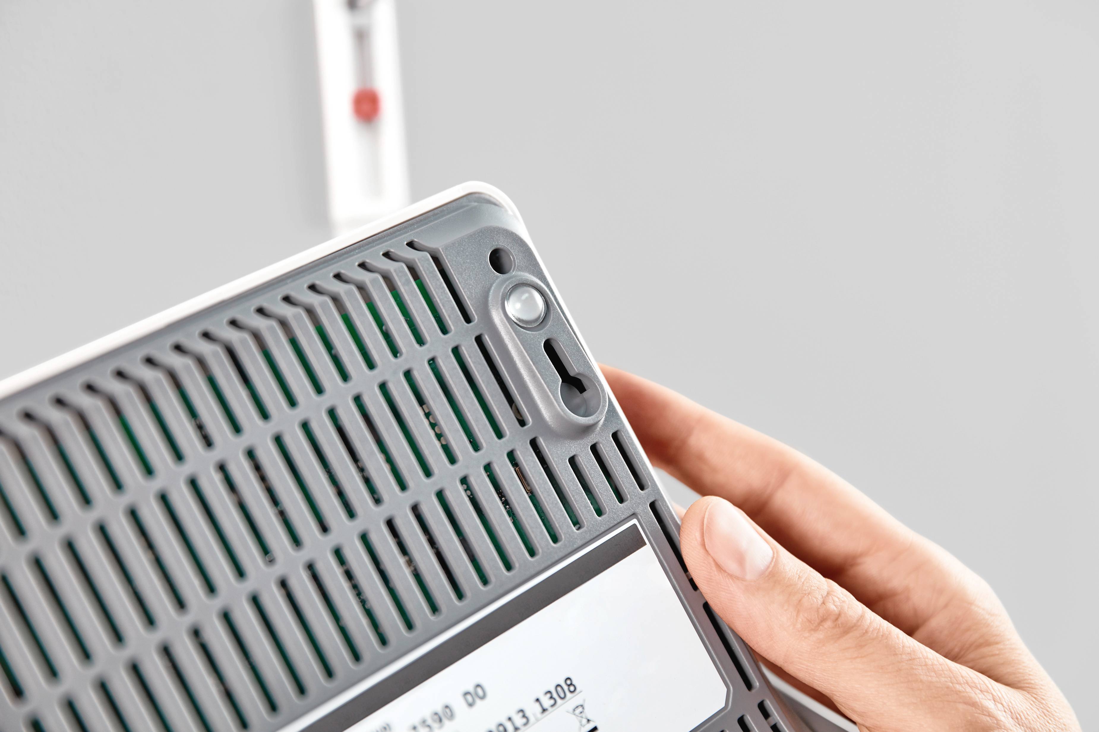 Eine Person hält einen grauen Gegenstand mit Lüftungsschlitzen und einem weißen Etikett. Hintergrund unscharf mit rotem Punkt.