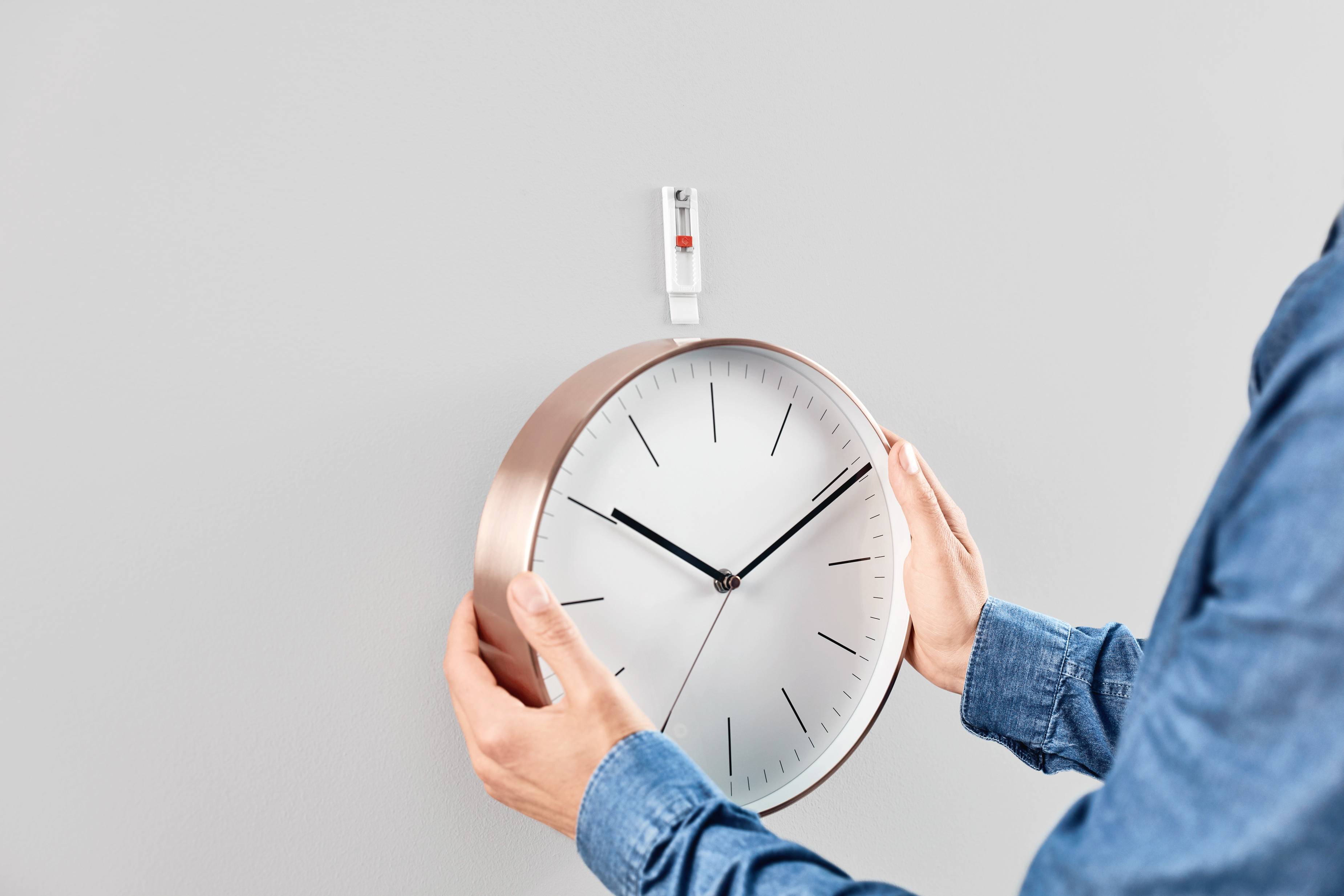 Eine Person hängt eine runde Wanduhr mit Kupferrahmen an einen Haken an der Wand. Die Uhr zeigt 10:10 Uhr.
