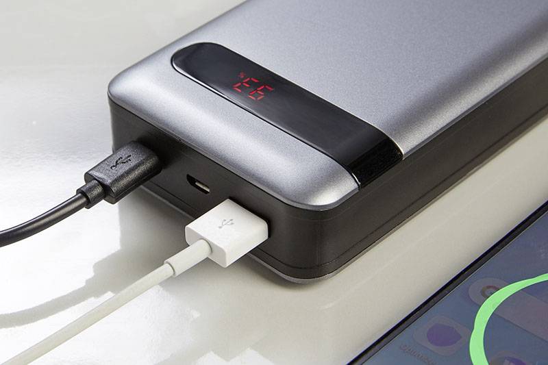 Eine Powerbank lädt ein Smartphone. Zwei Kabel sind angeschlossen; eins zeigt auf ein nahegelegenes Smartphone mit aktiviertem Display.