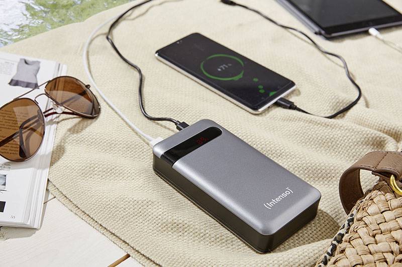 Tragbare Powerbank lädt Smartphone auf. Ein Tablet, eine Sonnenbrille und ein offenes Magazin liegen daneben auf einer Decke.