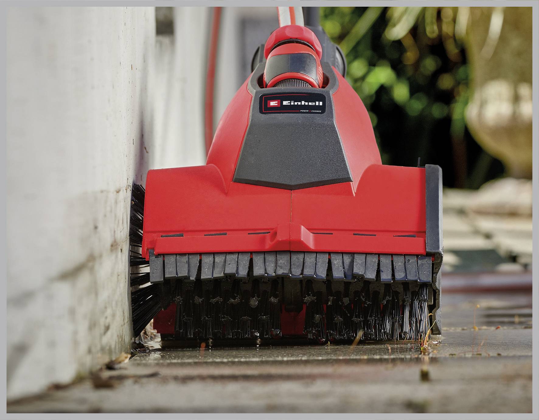 Ein roter elektrischer Bürstensauger reinigt eine Terrasse entlang einer Betonwand, entfernt Schmutz und Unkraut zwischen den Fugen.