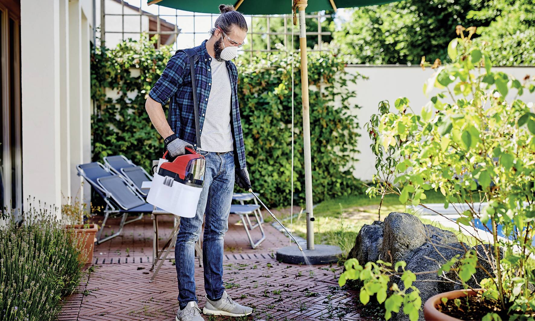 Person mit Schutzmaske und Handschuhen sprüht Pflanzen im Garten. Im Hintergrund sind Gartenmöbel und grüne Pflanzen zu sehen.
