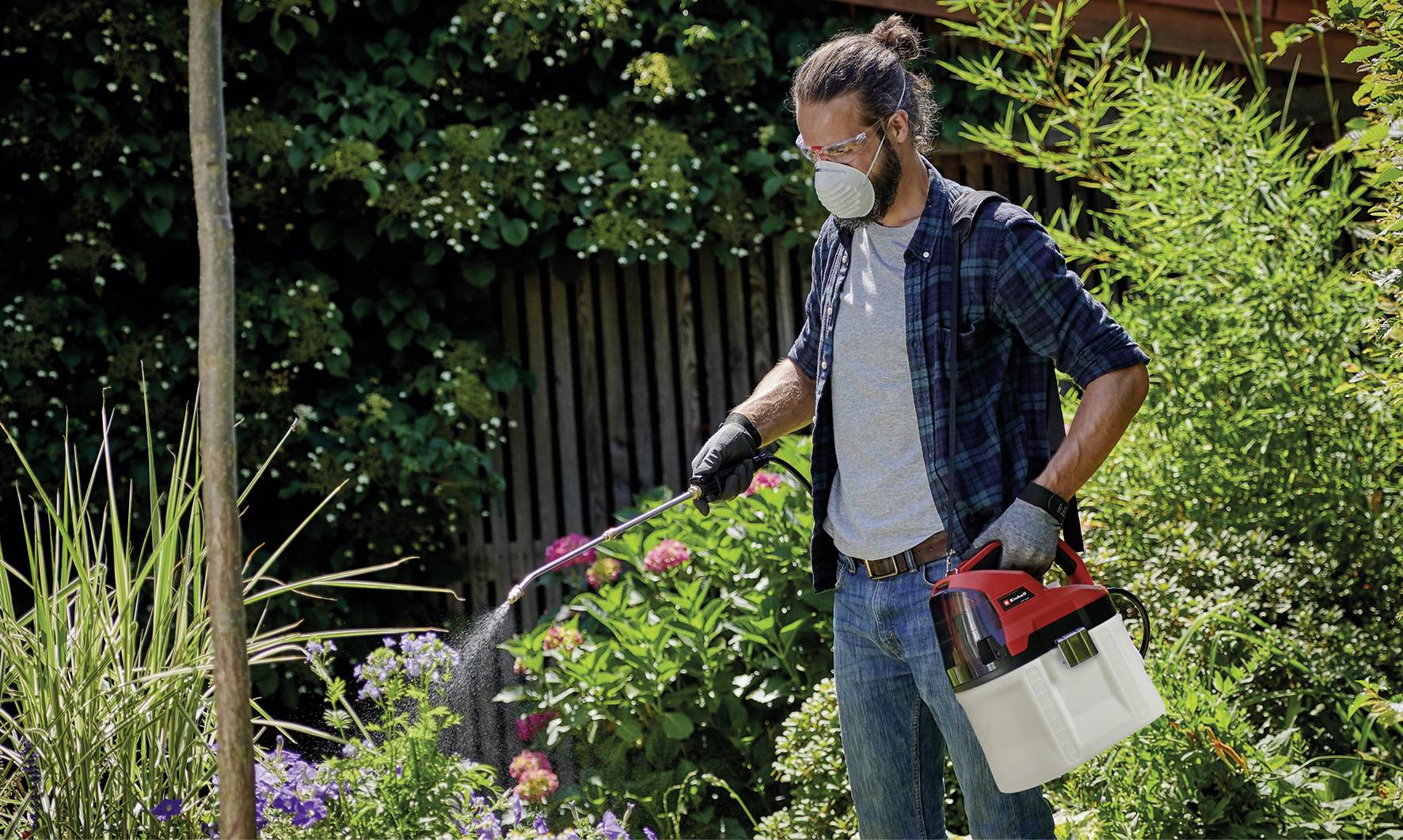 Person im Garten sprüht Pflanzen mit einem Gartensprühgerät. Trägt Schutzmaske, Handschuhe, Brille. Umgeben von Büschen und Blumen.