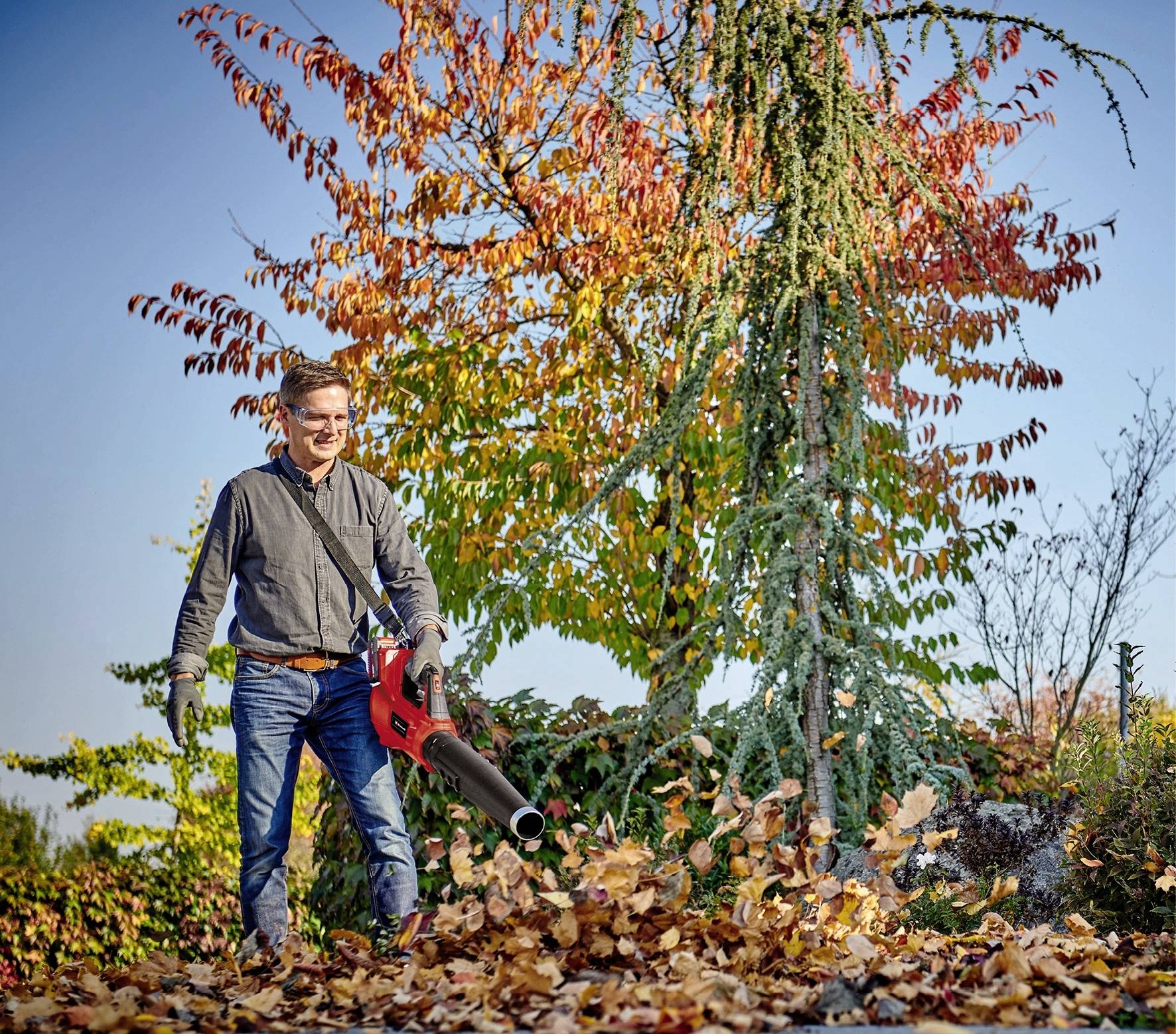 Ein Mann benutzt einen Laubbläser, um Herbstlaub in einem Garten wegzublasen. Im Hintergrund sind bunte Herbstbäume zu sehen.