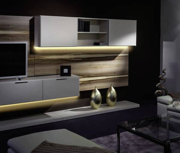 Wohnzimmer mit moderner, hängender Möbelwand in Holzoptik. LED-Beleuchtung, zwei goldene Vasen, Glas-Couchtisch und schwarzer Hintergrund.