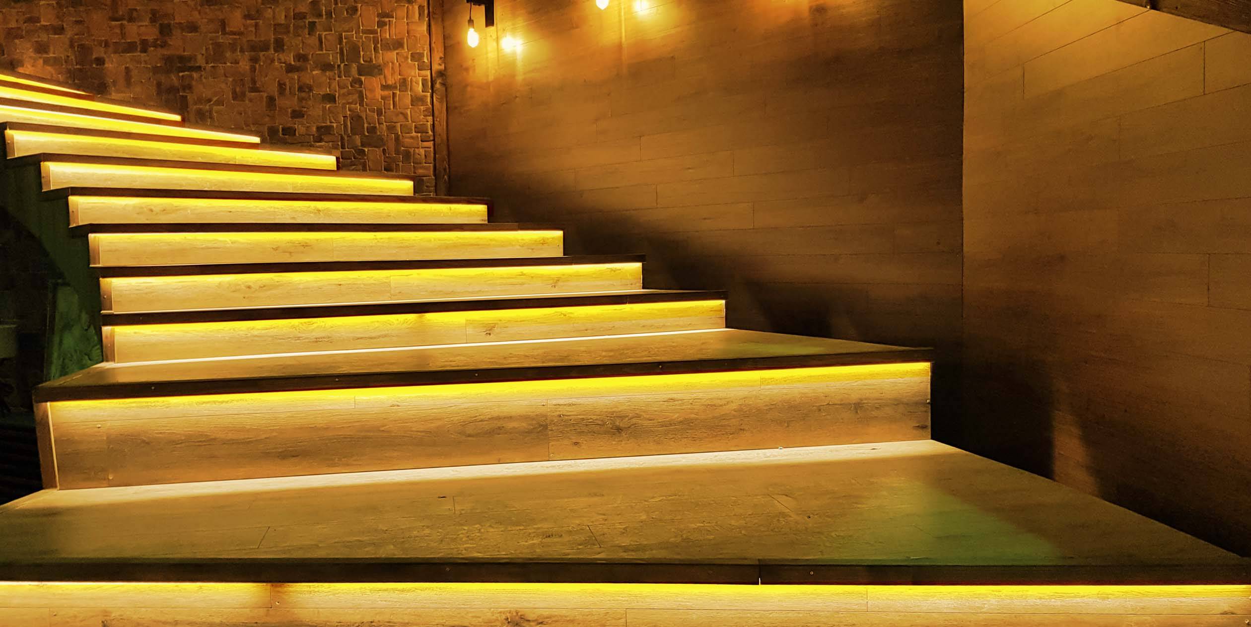 Treppe mit warmem Licht, das die Stufen hervorhebt, in einem gedämpft beleuchteten Raum mit Ziegel- und Holzwänden.