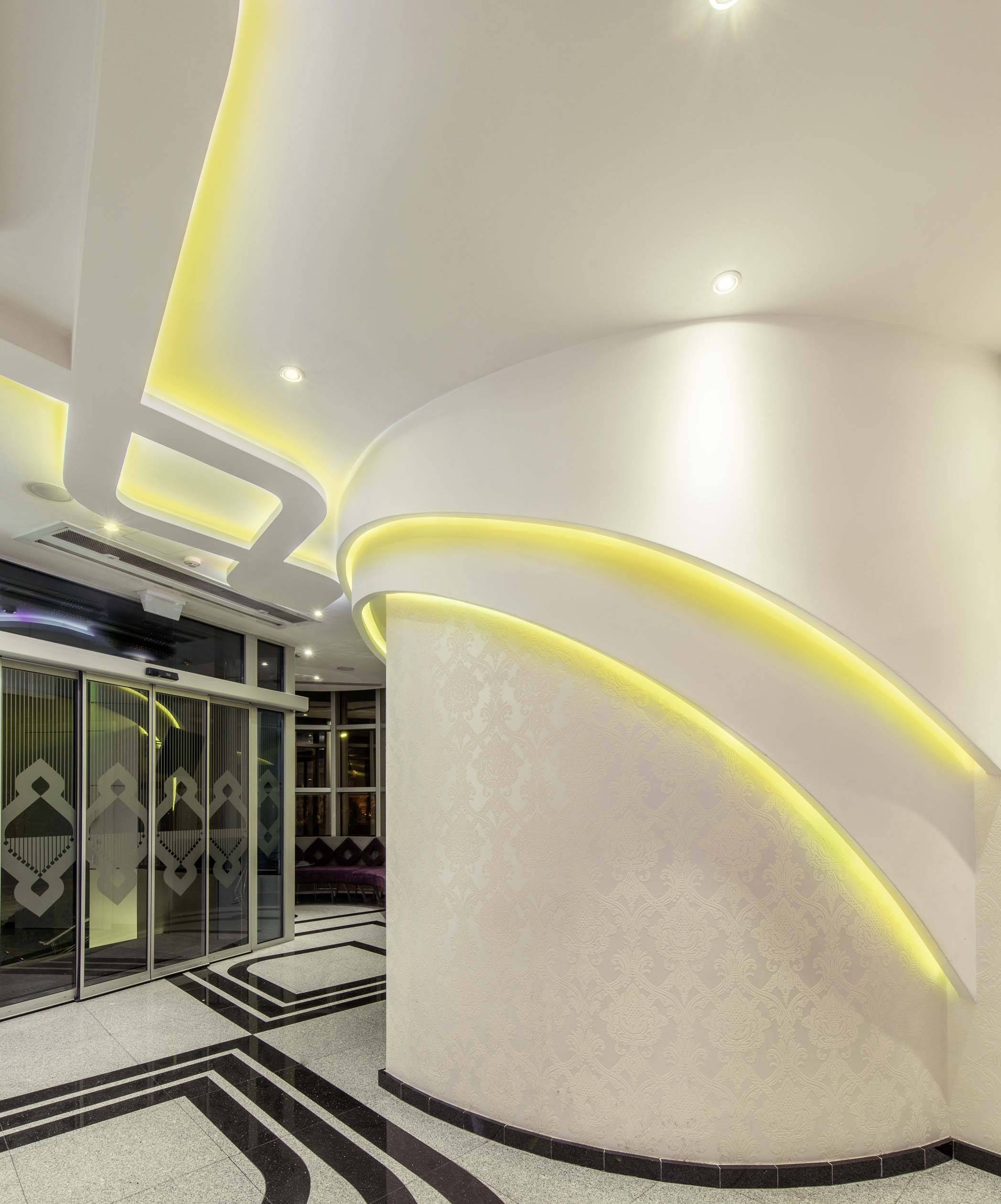 Modernes Foyer mit geschwungenen Wänden und gelben LED-Lichtern. Schwarz-weißes geometrisches Bodenmuster und Glastüren links.