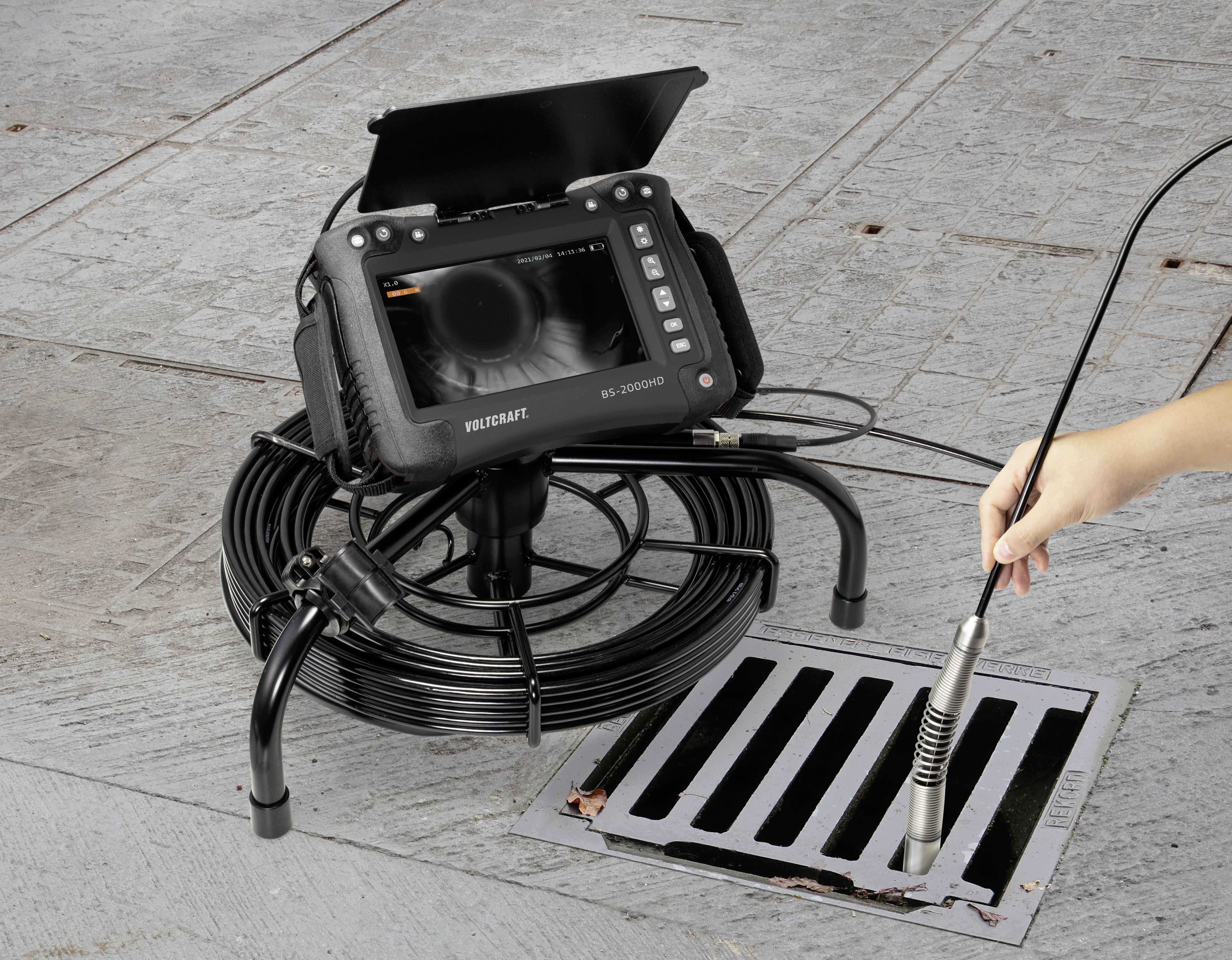 Eine Hand führt eine Kamera mit flexiblem Kabel in einen Straßenabfluss ein, während ein Überwachungsmonitor das Innere zeigt.