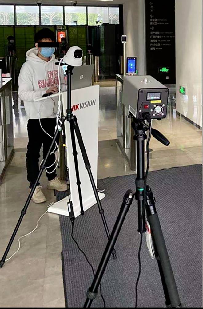 Eine Person mit Maske steht hinter einem Laptop auf einem Stativ. Ein weiteres Stativ mit einem Wärmebildgerät im Vordergrund.