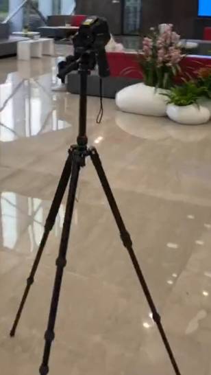 Dreibeinstativ mit montierter Kamera in modernem Raum mit glänzendem Boden und dekorativen Pflanzen im Hintergrund.