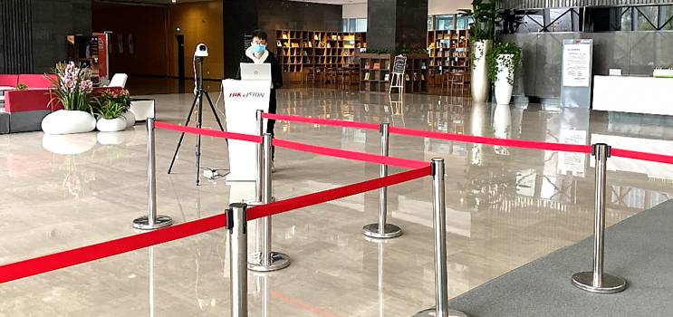 Empfangsbereich mit abgesperrtem Zugang durch rote Bänder, Scanner auf Stativ, Mitarbeiter mit Maske am Stand. Bücherregal im Hintergrund.