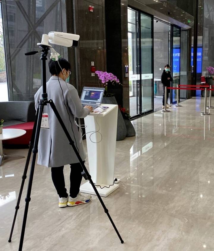 Person mit Laptop an weißem Podest in einer Lobby, technologisches Gerät auf Stativ daneben, weiterer Mensch im Hintergrund.