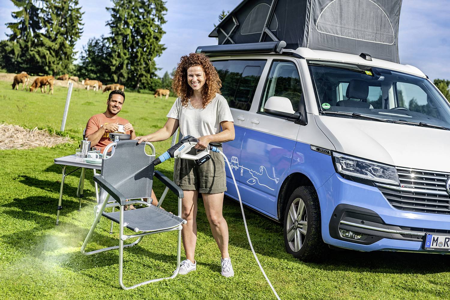 Ein Mann sitzt auf einem Campingstuhl und trinkt Kaffee neben einem blauen Wohnmobil; im Hintergrund weiden Kühe auf einer Wiese.