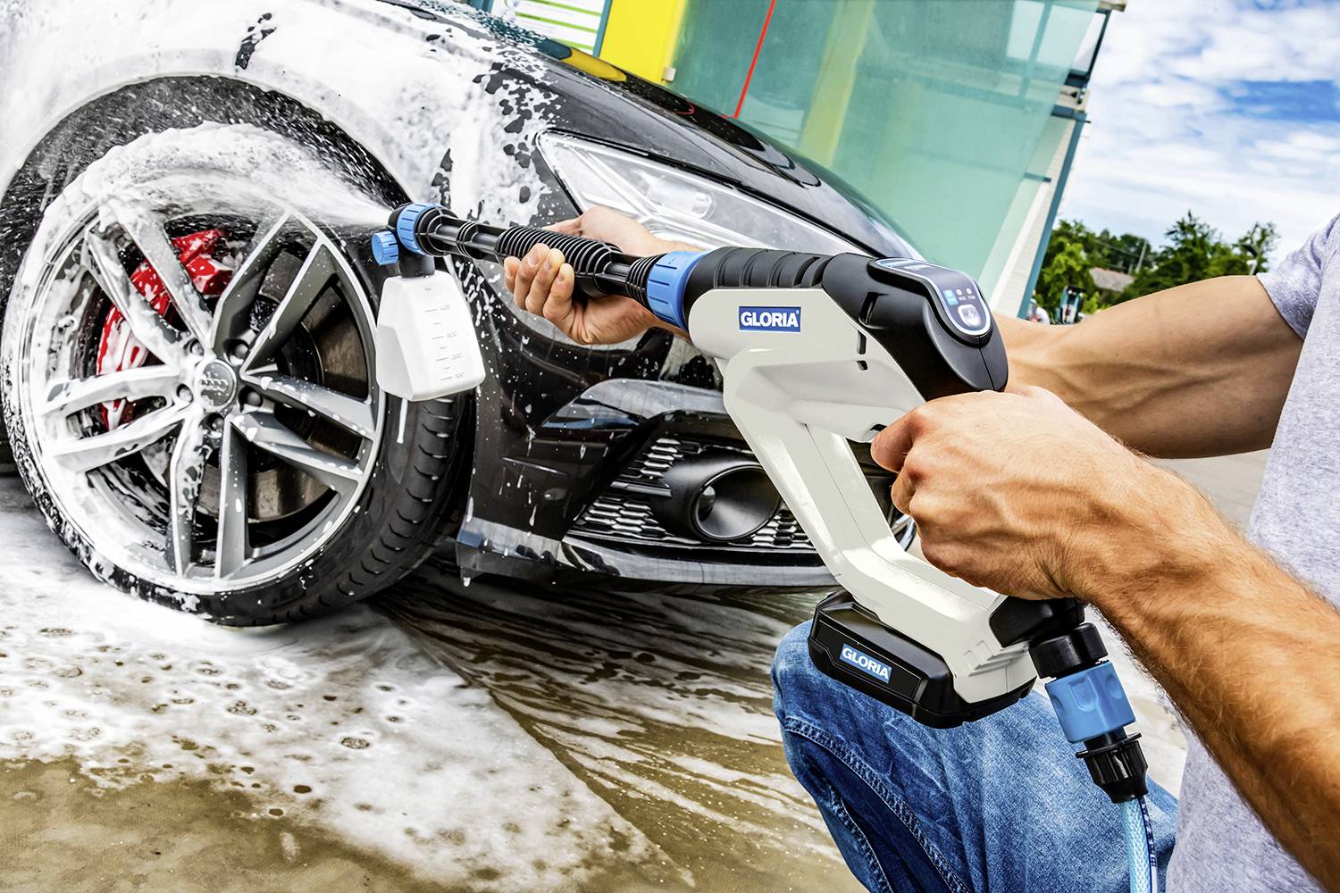 Eine Person wäscht ein Auto mit einem Hochdruckreinigungsgerät, sprüht Wasser auf das hintere Rad und Schaum bedeckt das Auto.