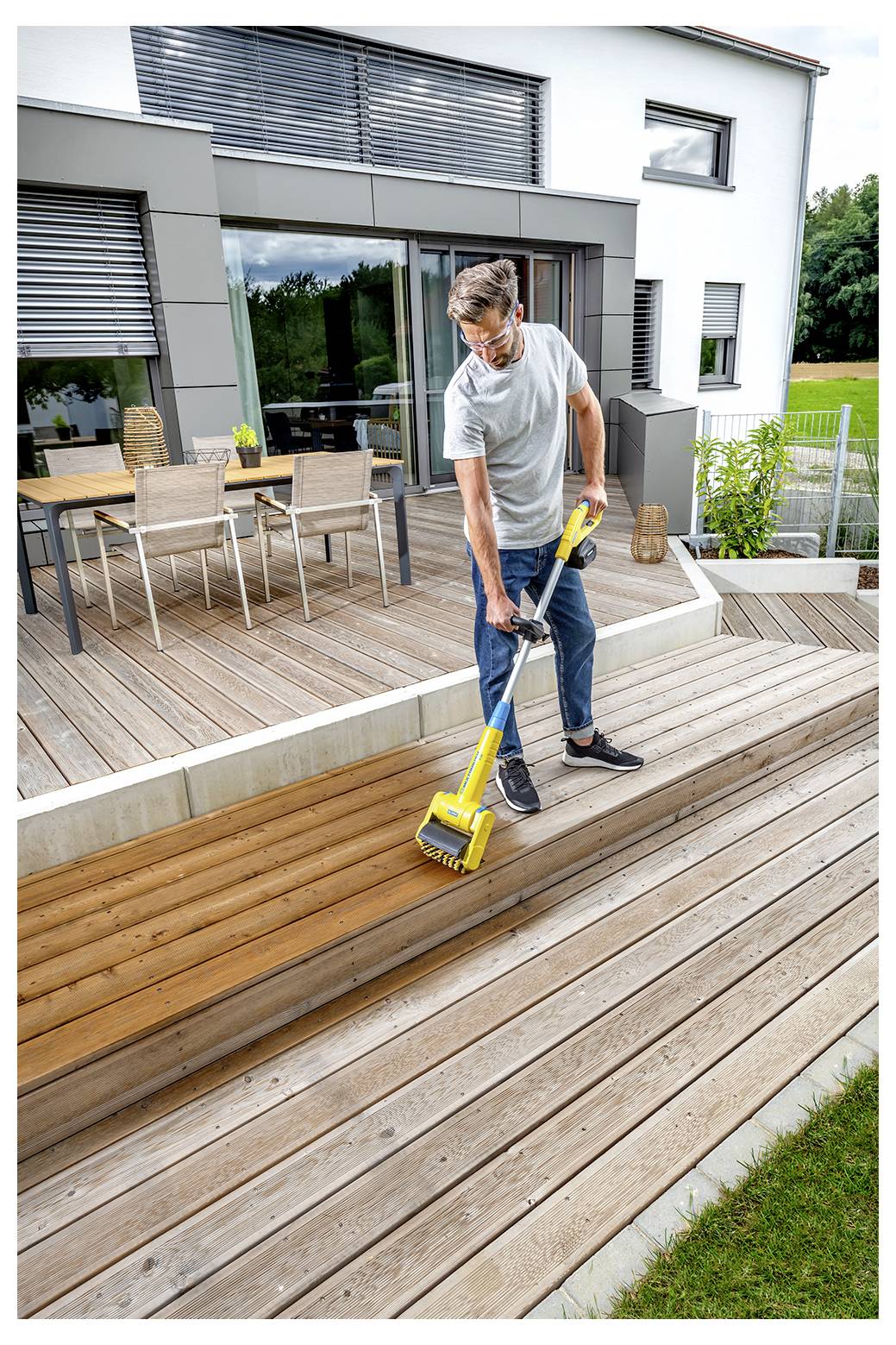 Ein Mann reinigt ein Holzdeck mit einer gelben Elektrobürste vor einem modernen Haus, mit Terrassenmöbeln im Hintergrund.