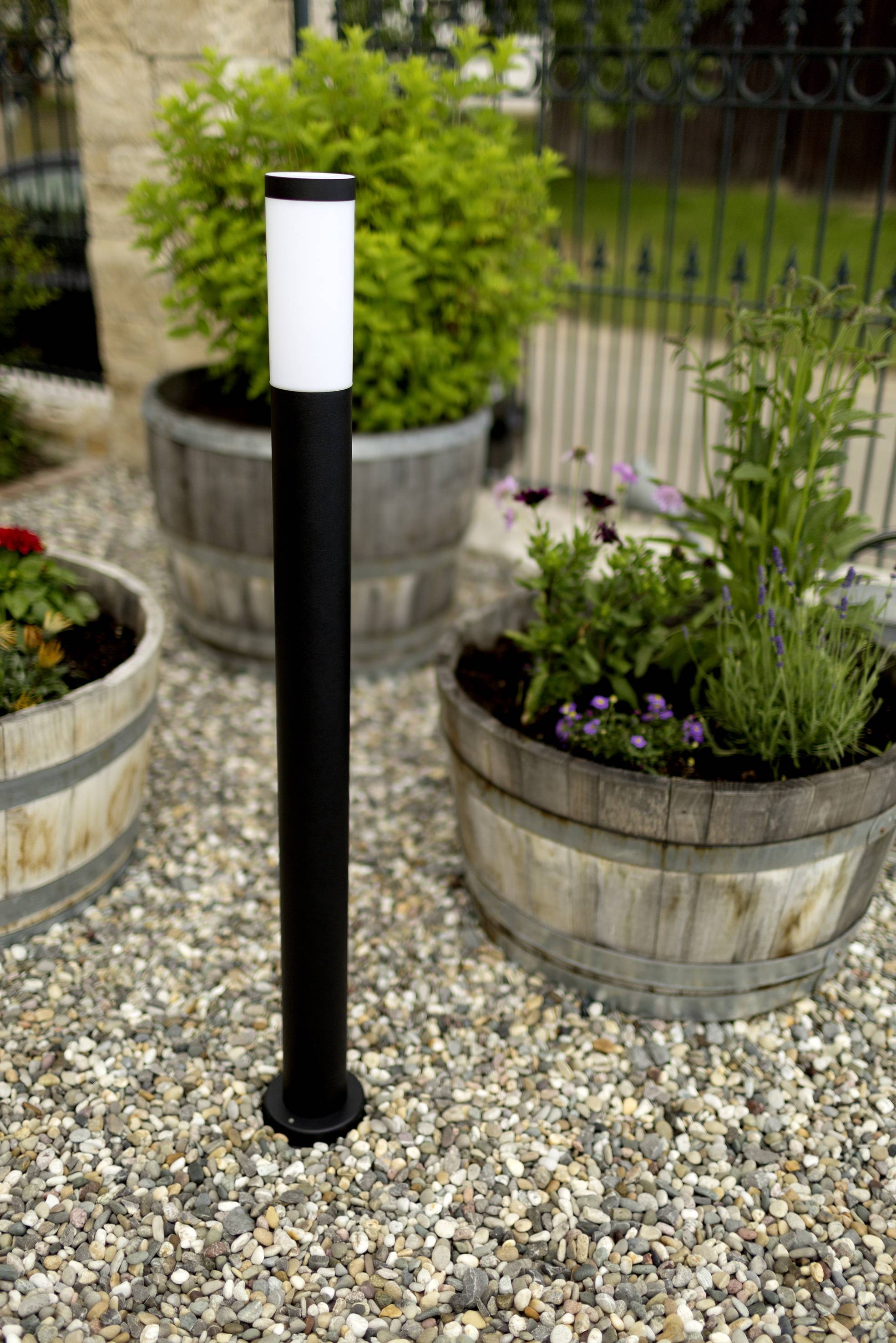 Schwarze Wegleuchte mit weißem, zylindrischem Kopf steht auf einem Kiesweg, flankiert von Holzkübeln mit grünem und blühendem Pflanzen.