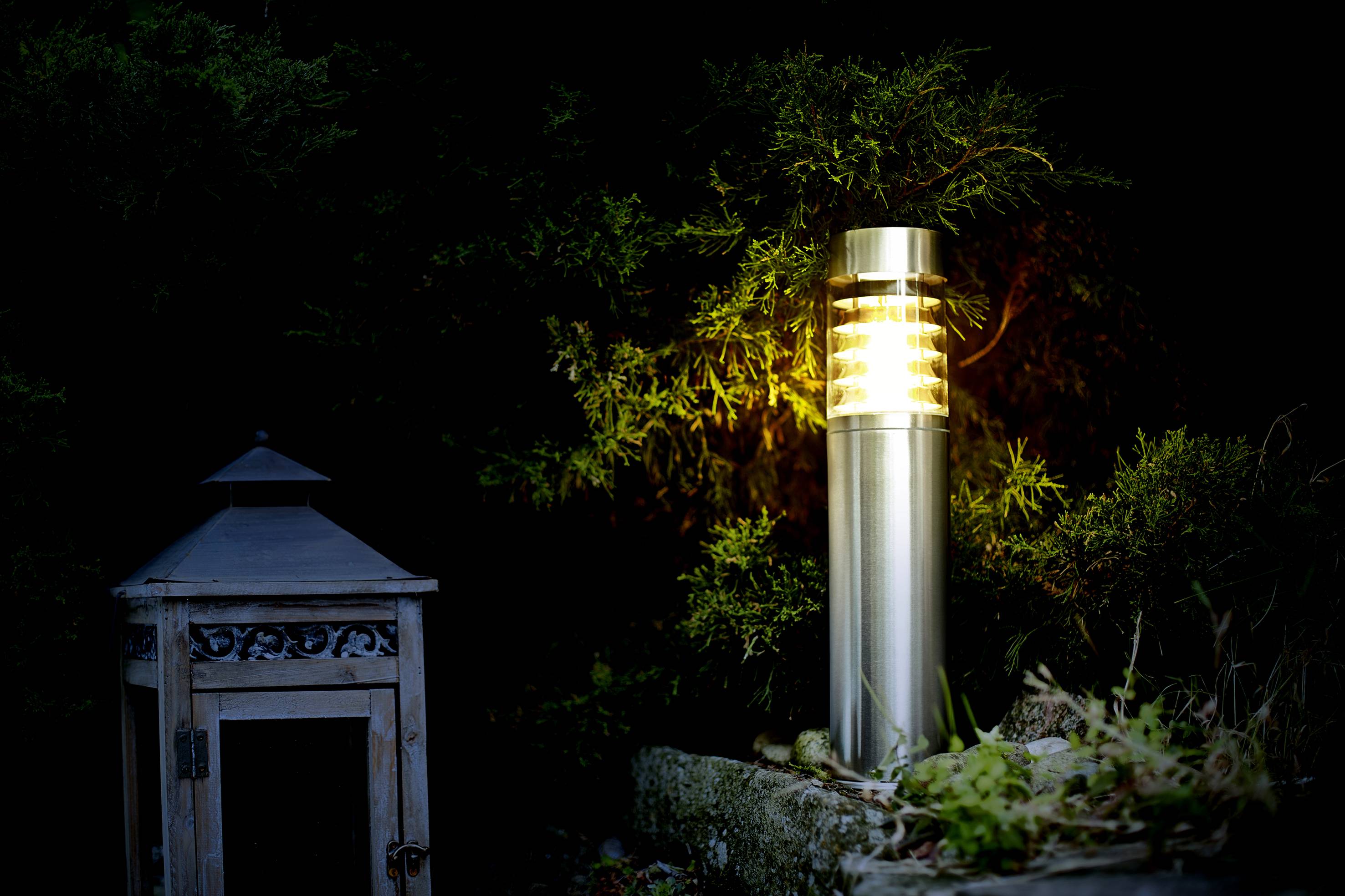 Gartenlaterne aus Metall leuchtet in der Nacht neben einem alten, hölzernen Laternengehäuse, umgeben von dunklem Laub.