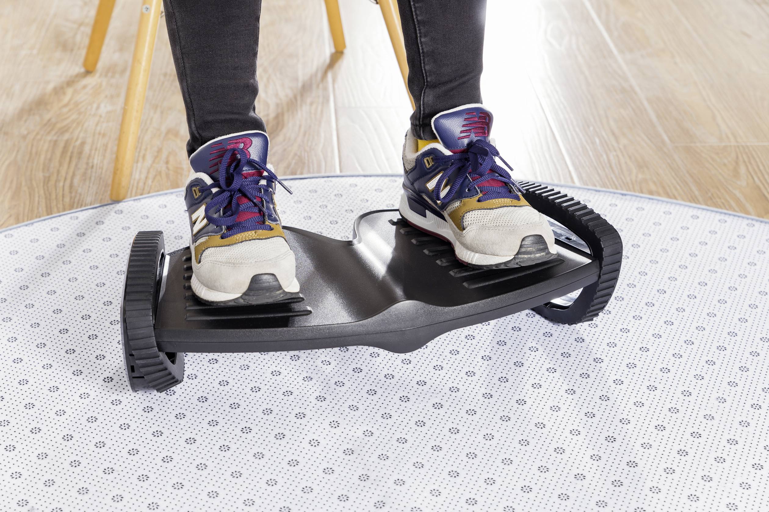 Zwei Füße in Turnschuhen stehen auf einem schwarzen Wippbrett mit geriffelten Rädern, auf hellem Holzboden und Teppich.