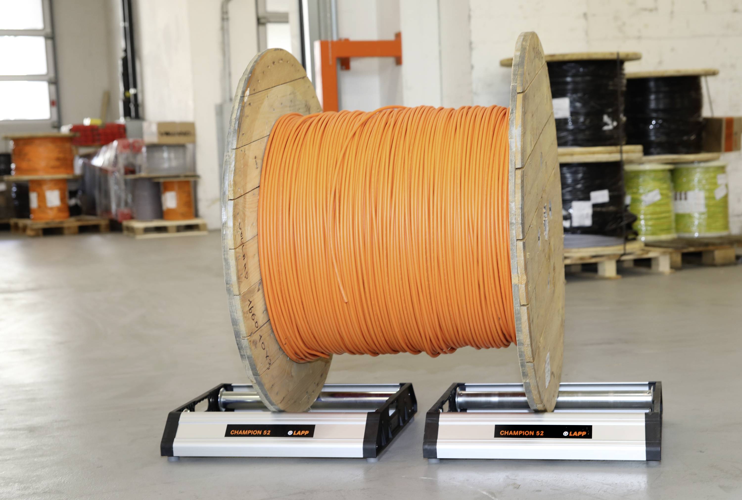Eine große Kabeltrommel mit orangefarbenem Kabel steht in einer Lagerhalle. Im Hintergrund sind weitere Kabeltrommeln auf Paletten sichtbar.