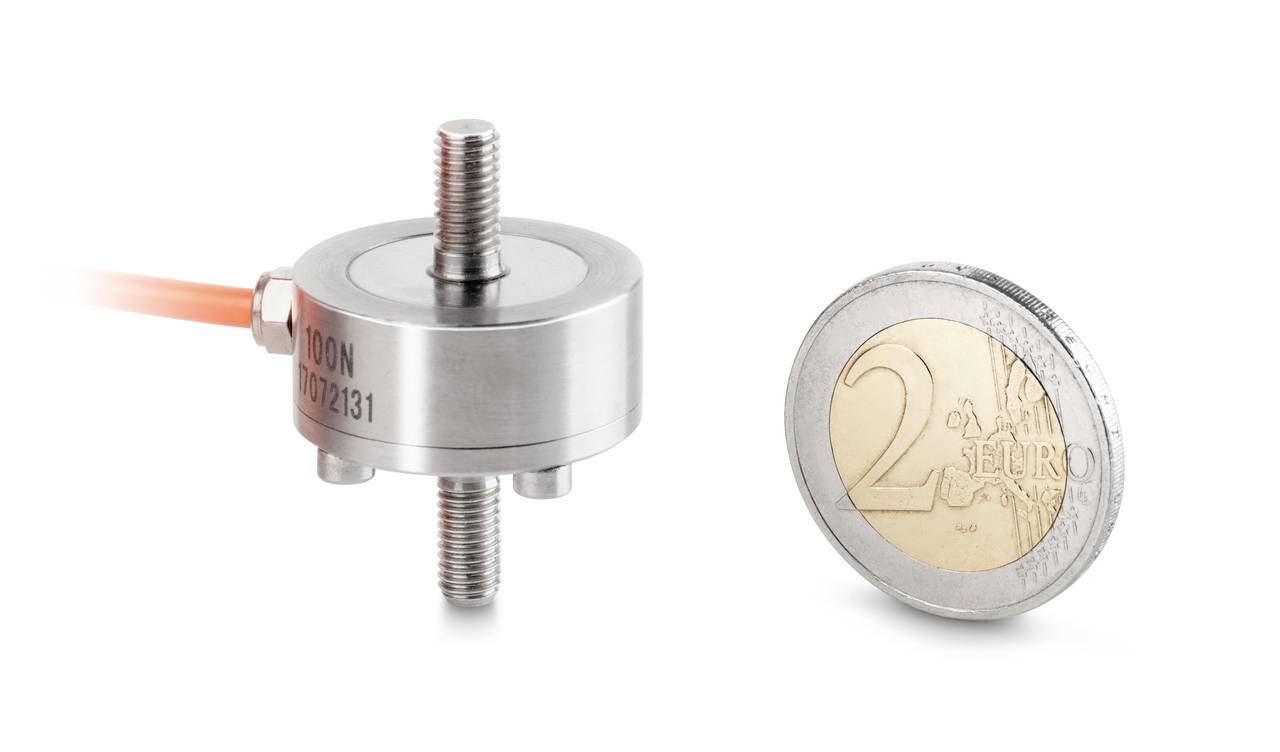 Kraftsensor neben einer 2-Euro-Münze zum Größenvergleich. Der Sensor hat eine runde, kompakte Form mit einem Metallgehäuse und Schraubverbindungen.