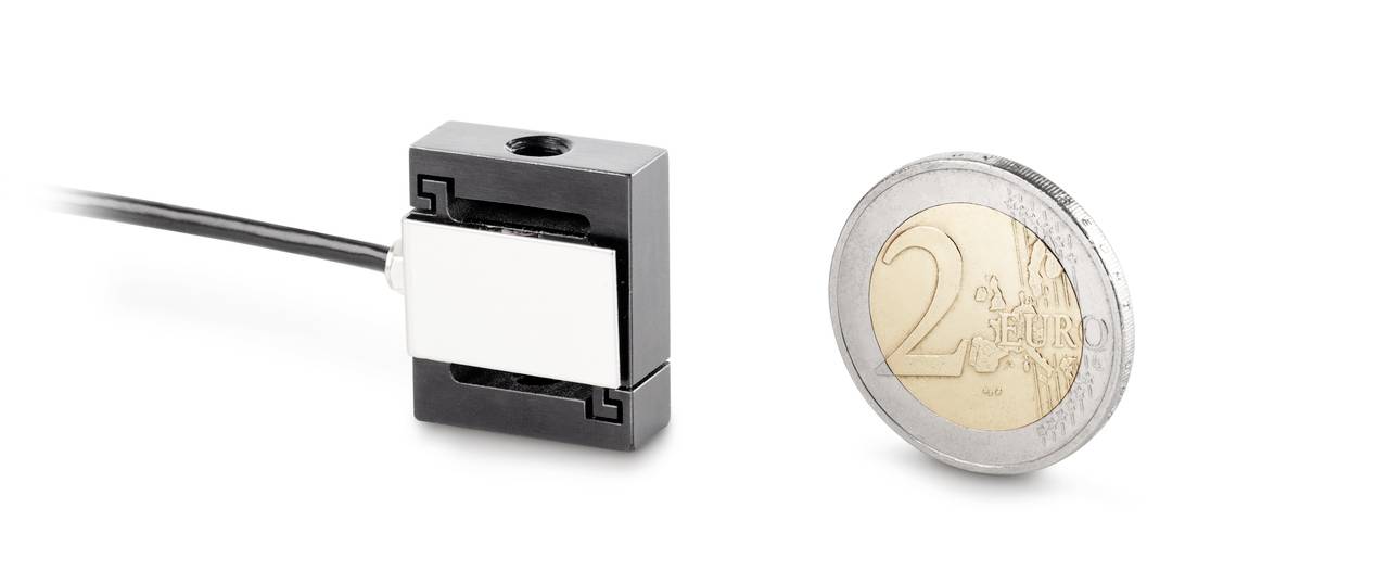 Ein kleines elektrisches Bauteil neben einer 2-Euro-Münze, die der Größenvergleich zeigt. Das Bauteil hat ein schwarzes Gehäuse und ein Kabel.