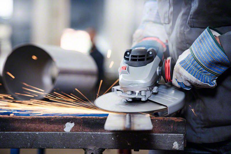 Person schleift Metall mit einem Winkelschleifer, Funken sprühen; trägt Schutzhandschuhe. Arbeitsplatz in einer Werkstatt.