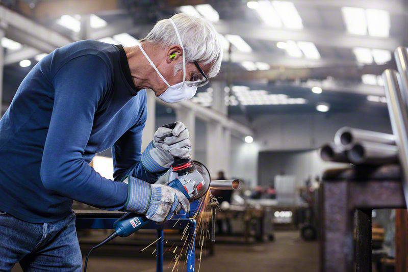 Ein Mann in Arbeitskleidung und Maske schleift ein Metallrohr in einer Werkstatt. Funken fliegen vom Werkzeug.