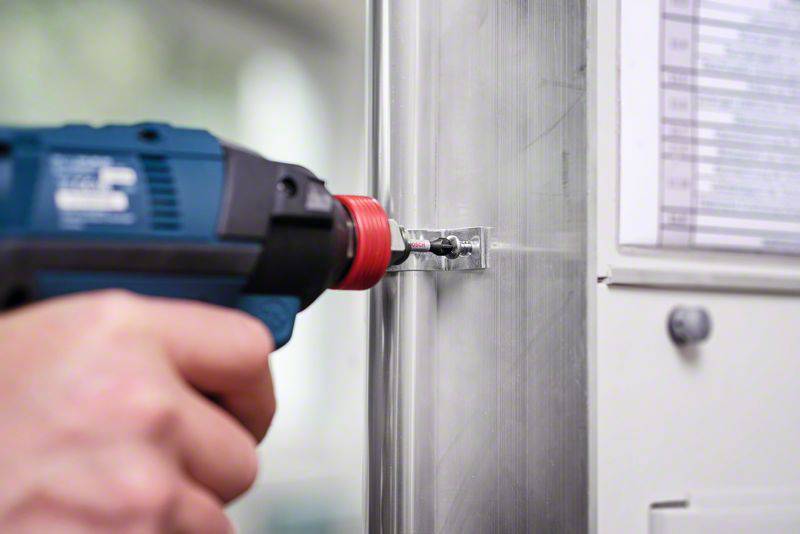 Eine Person verwendet einen blauen Akkuschrauber, um eine Schraube in ein Metallgehäuse zu befestigen. Im Hintergrund sind verschwommene technische Geräte sichtbar.